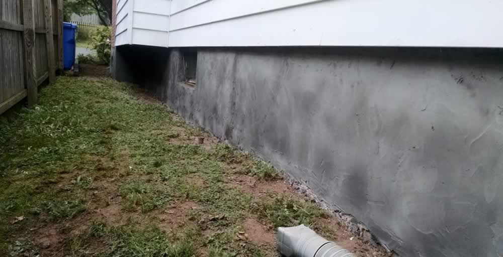 Narrow grassy side yard beside a house with a gray foundation wall and a wooden fence on the left.