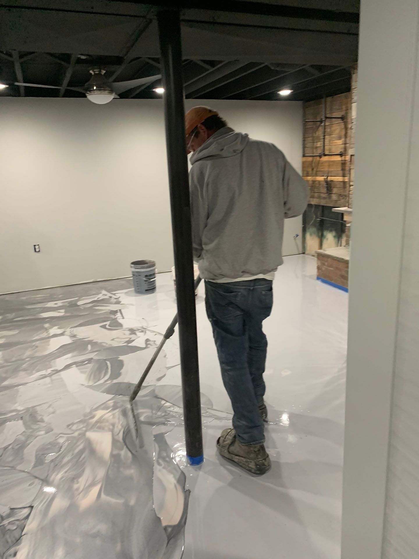 Worker painting an indoor room with a roller, floor covered in plastic sheeting, walls partially finished