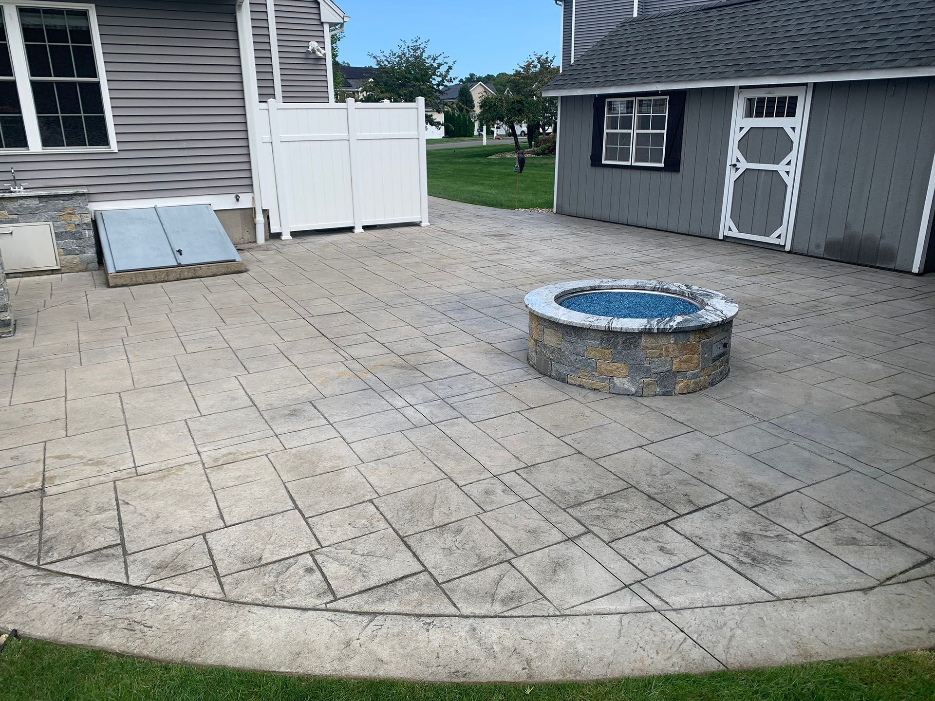 Backyard patio with paver stones, a stone fire pit, and small buildings/sheds nearby.