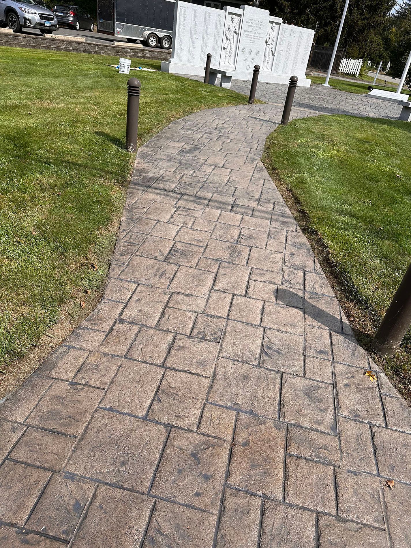 Curved brick walkway through a grassy area, with short black posts and a white structure ahead.