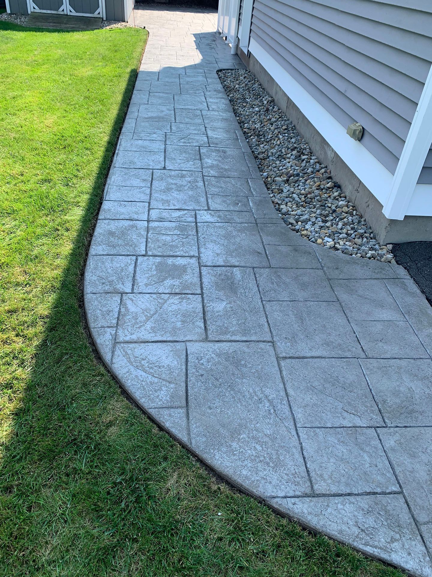 Curved stone paver walkway beside a house, bordered by grass and a gravel strip