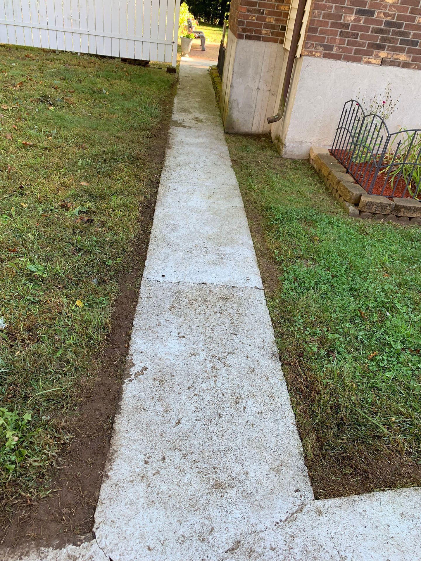 Concrete walkway between grass strips beside a white house and fence, leading to a sunlit gate