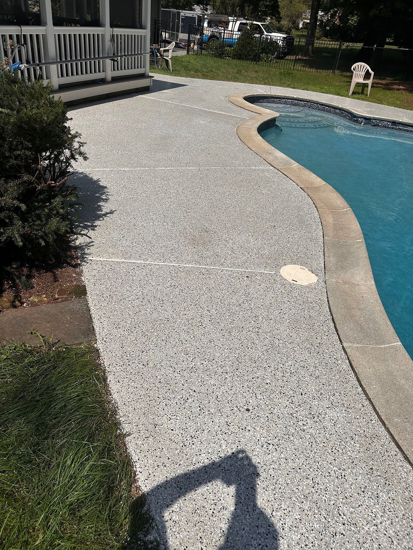 Pebble-finished pool deck beside a curved swimming pool, with white railing and a chair in the background