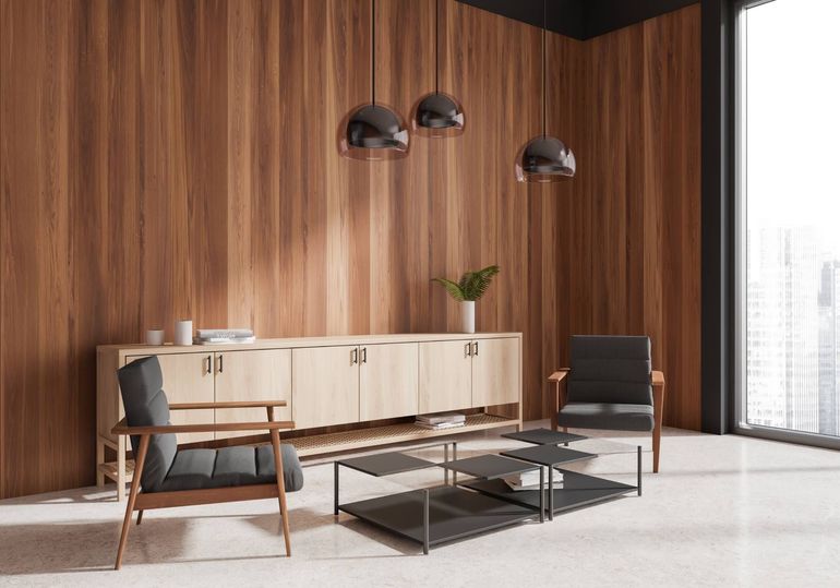 Modern lounge with wood-paneled wall, low cabinet, black chairs, and glass coffee tables by a window.