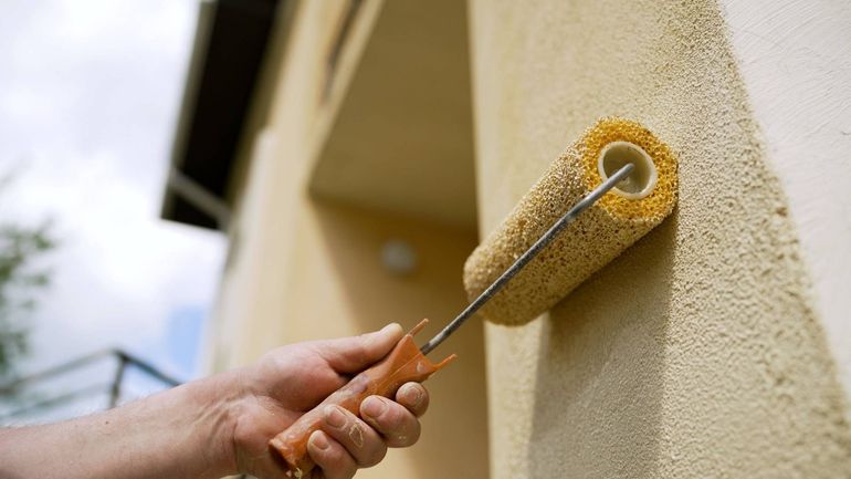 Hand using a paint roller to apply yellow paint to a house wall.