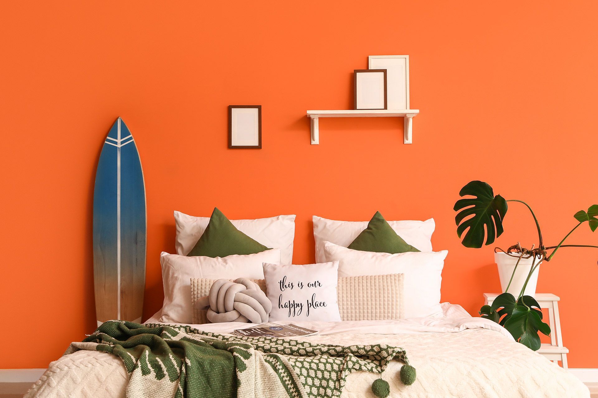 Cozy bed with white and green pillows against a bright orange wall, surfboard and plants in a stylish bedroom.
