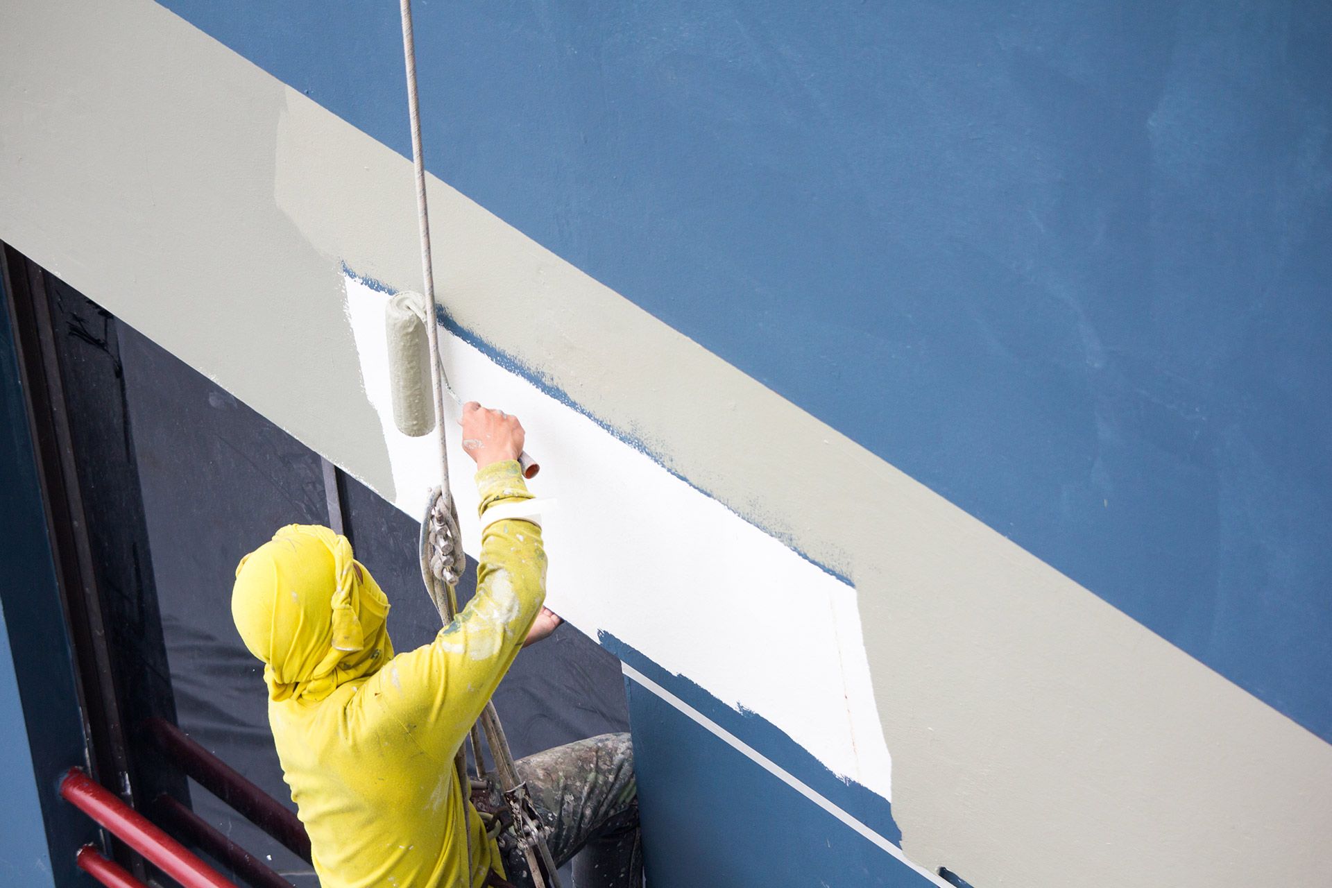 Person in yellow rain jacket painting a wall with a roller and brush on blue-and-white interior surface