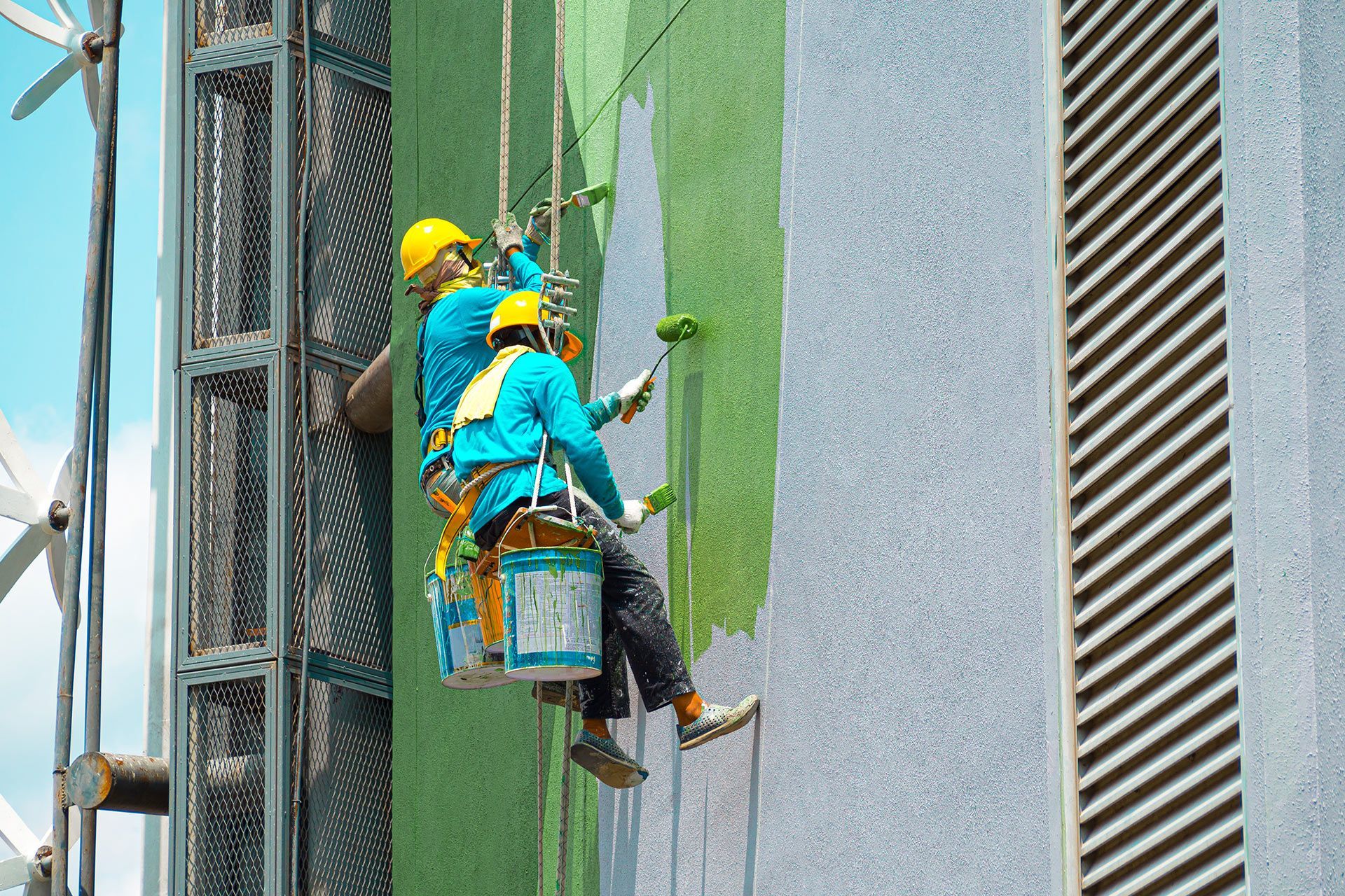 Two workers suspended on a building facade, painting a wall green from a bucket lift