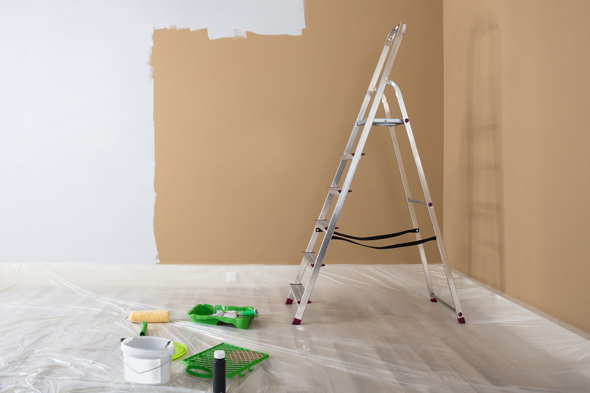 Painting supplies and ladder in a room with a beige wall being painted.