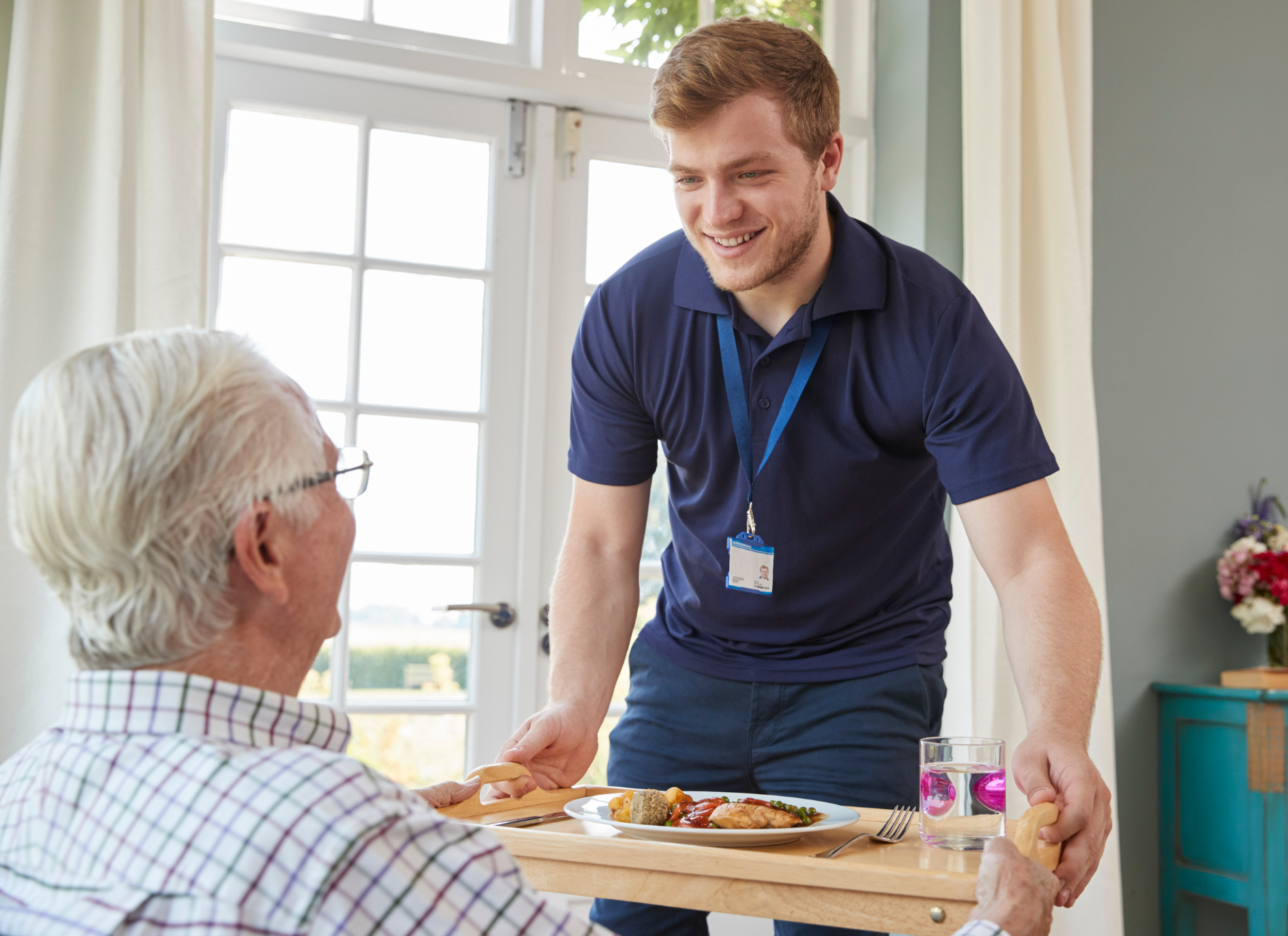 Caregiver serving a meal to an older adult at a table. Both smile, inside a home, bright room, sunny.