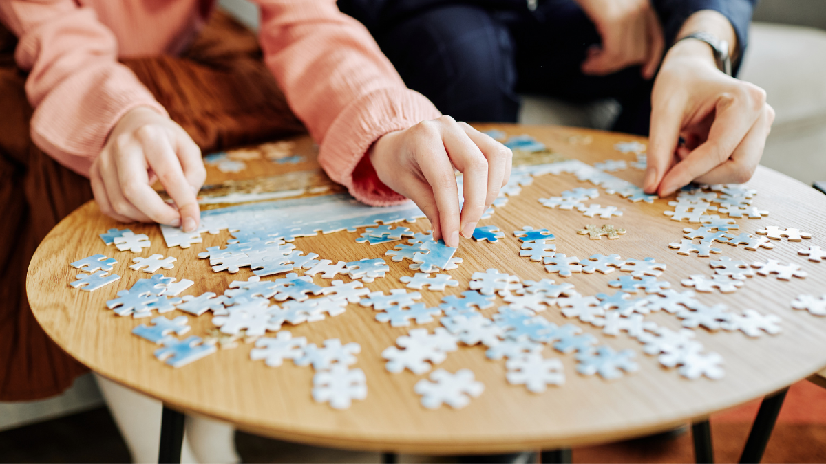 Two people assembling a jigsaw puzzle on a small wooden table; blue and white pieces.
