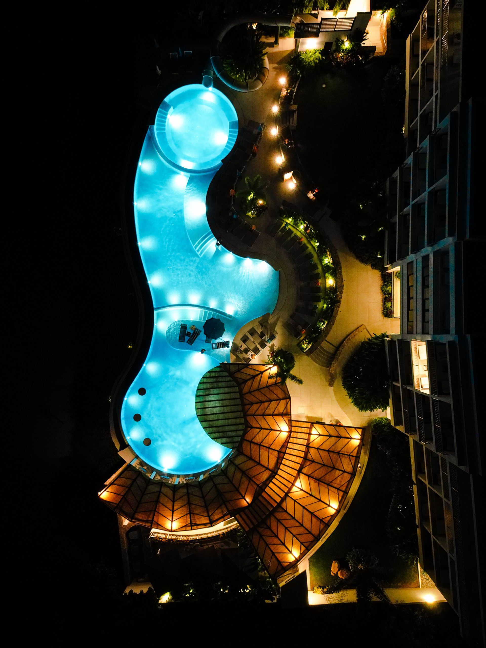 Aerial nighttime view of a lit blue swimming pool beside a warmly lit resort courtyard