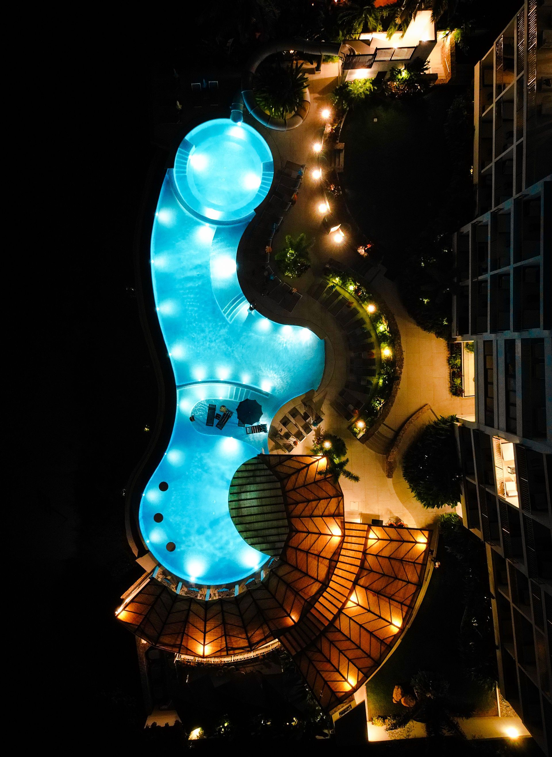 Aerial view of a lit blue kidney-shaped pool beside palm trees and a winding walkway at night