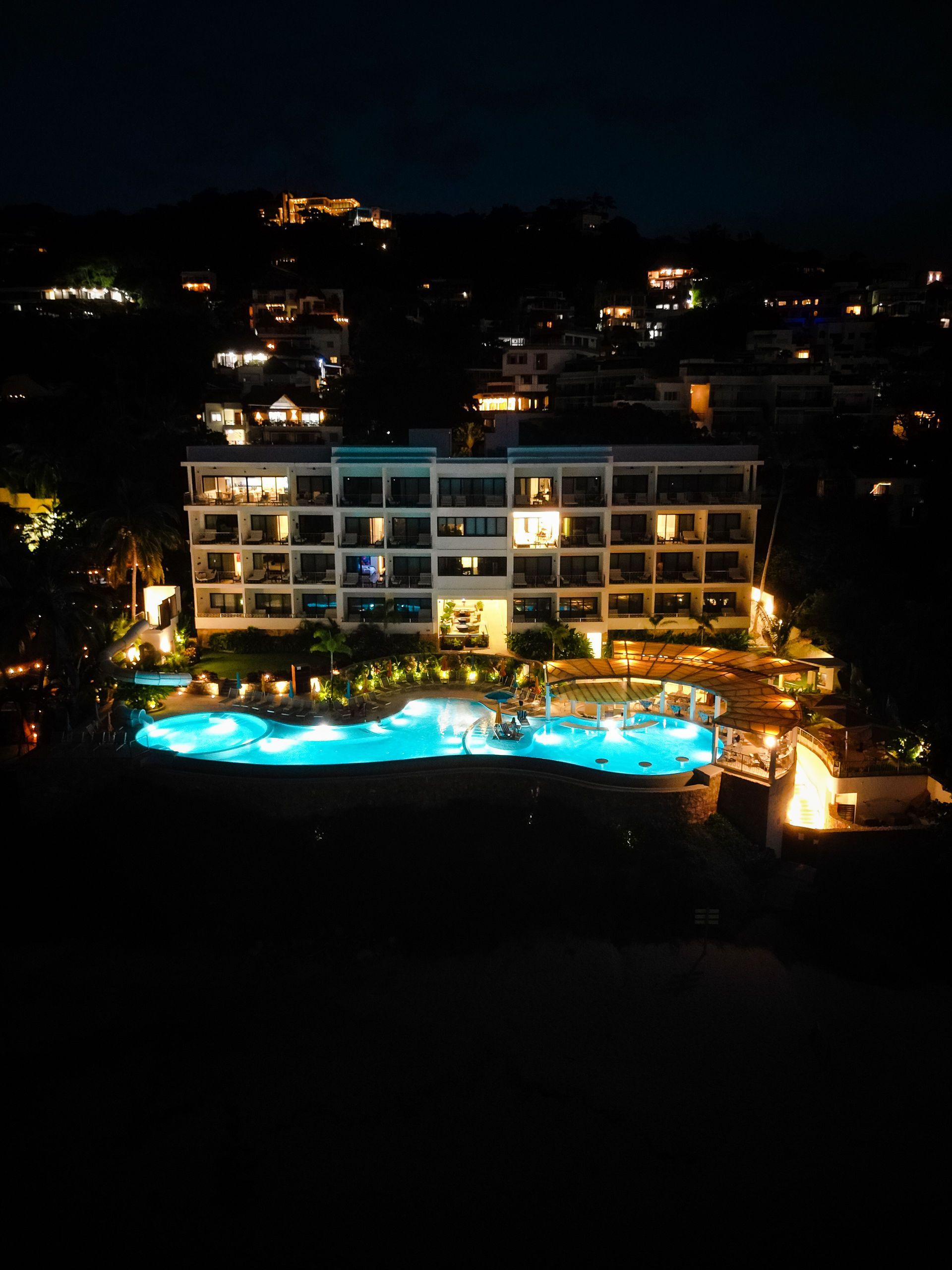 Nighttime resort hotel with glowing blue pool and lit balconies on a dark hillside