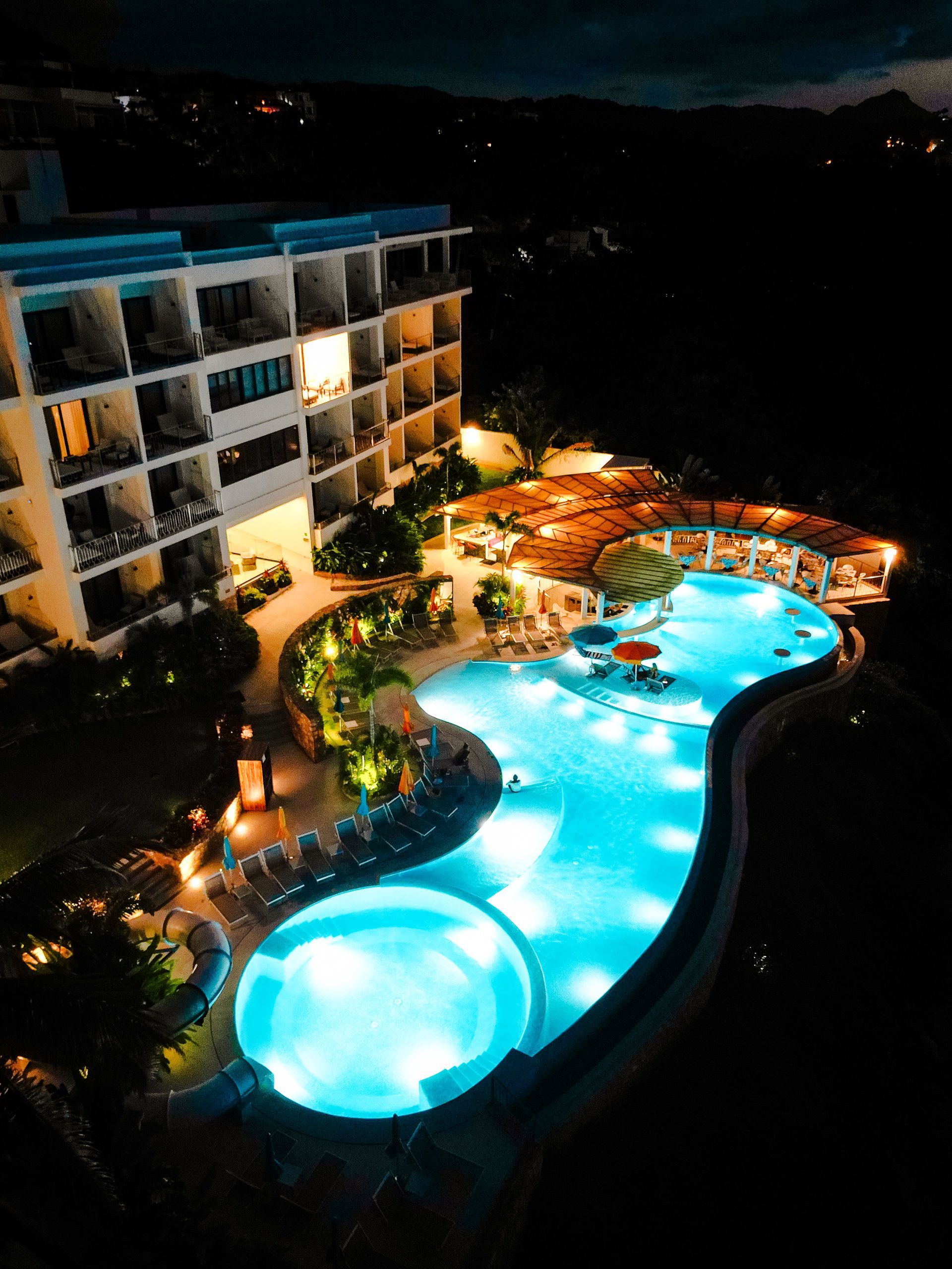 Nighttime resort pool lit blue beside a multi-story hotel with warm balcony lights