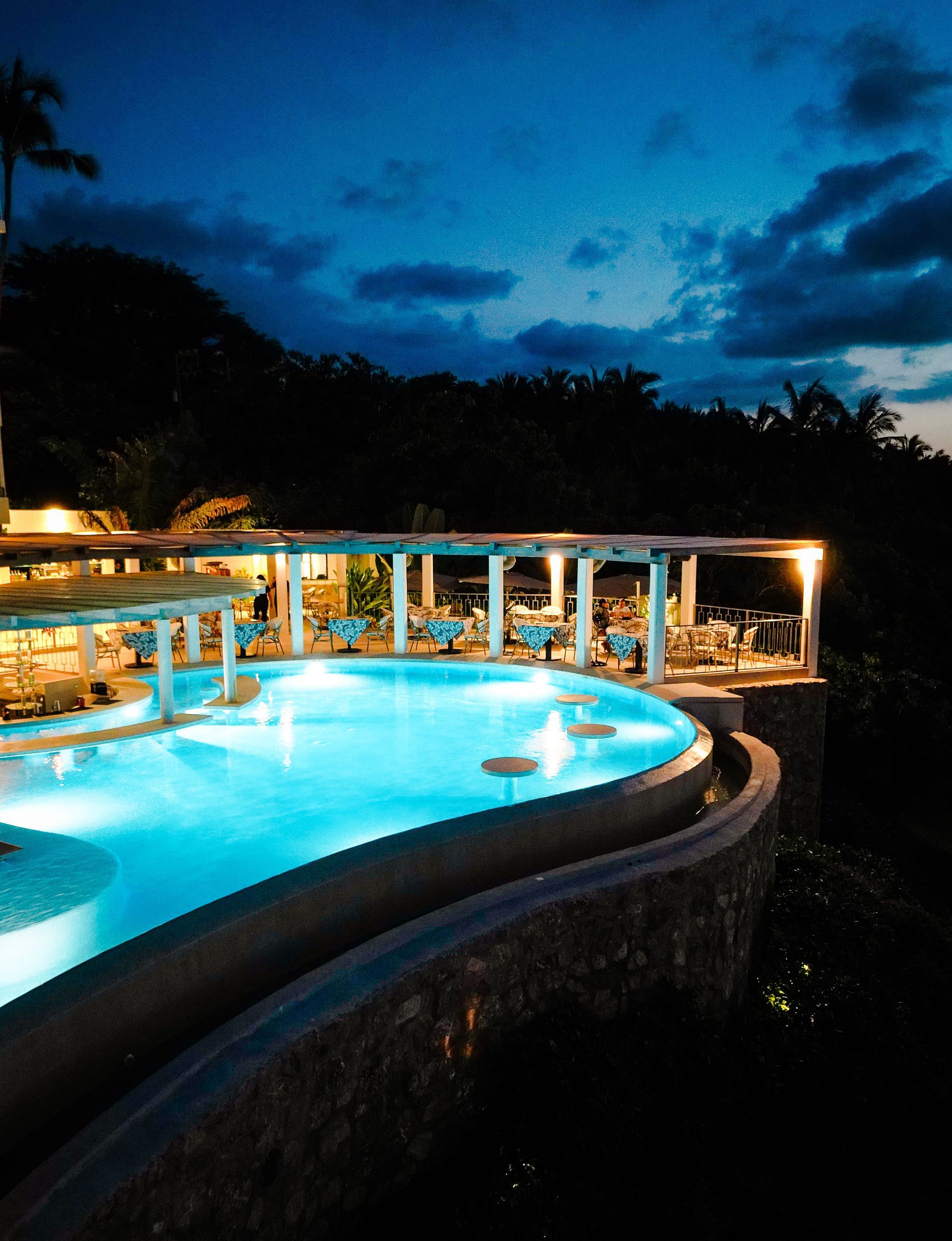 Illuminated resort pool at dusk with turquoise water, white columns, and dark trees under a blue evening sky