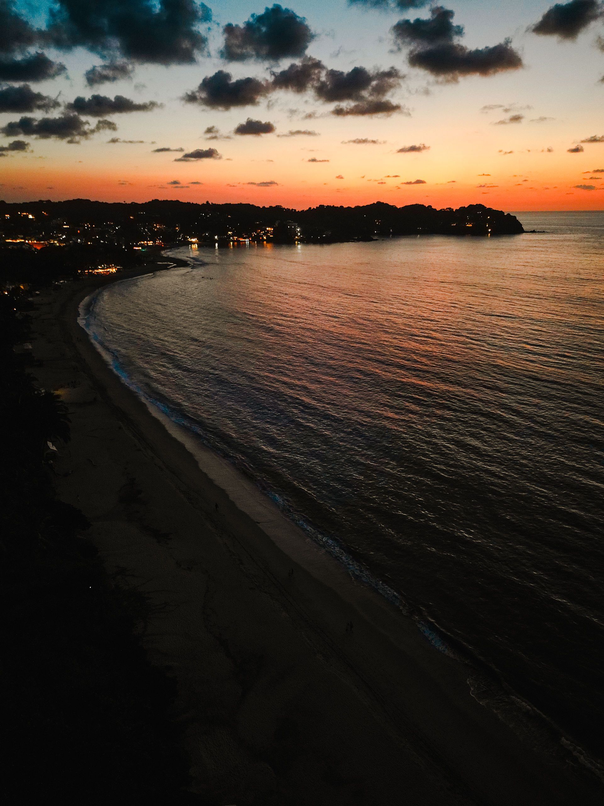 Sunset over a calm beach with glowing shoreline lights and dark waves under a pink-orange sky