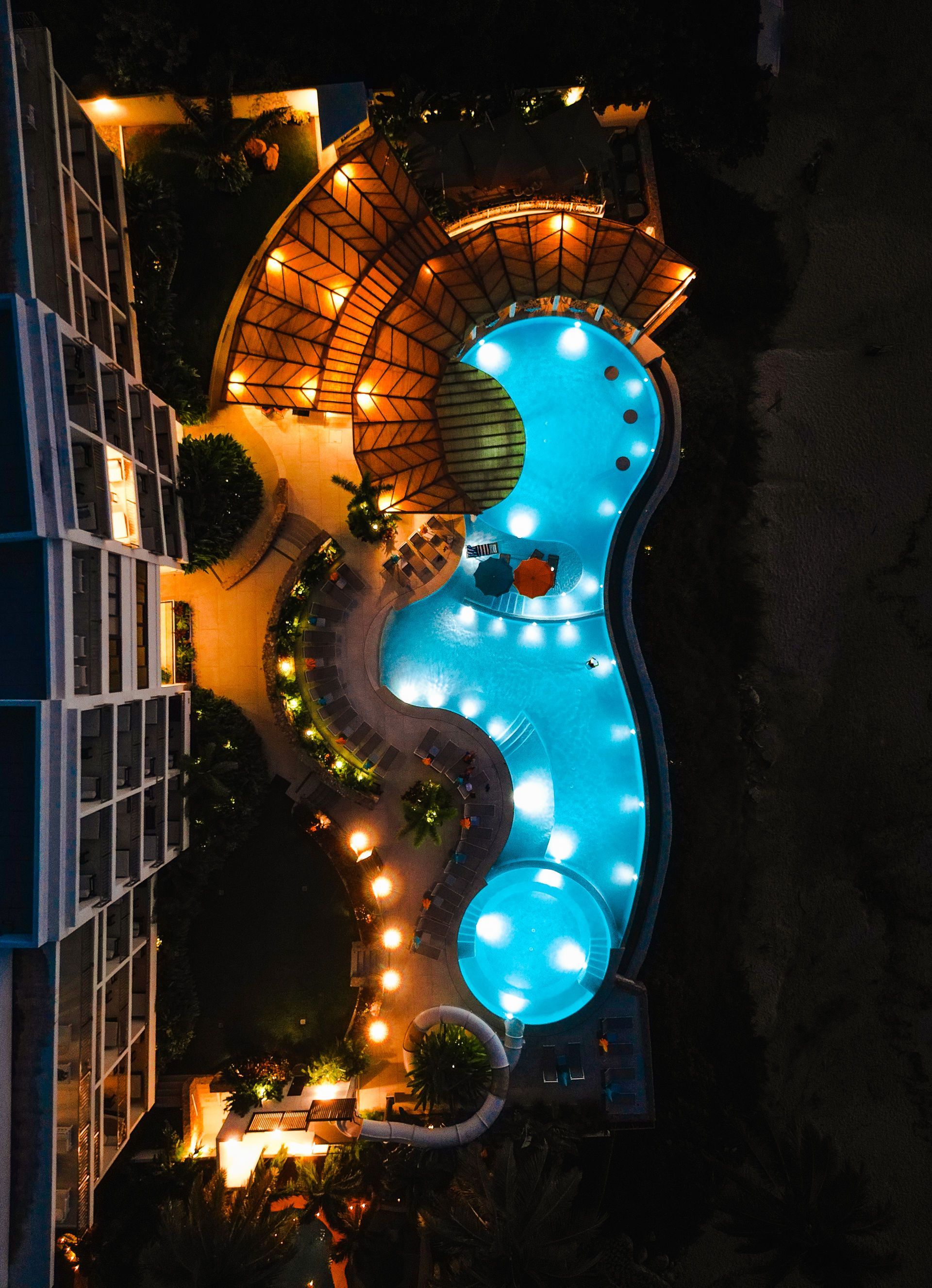 Aerial view of a lit resort pool at night beside a building, with a curving blue swimming pool and warm lights.