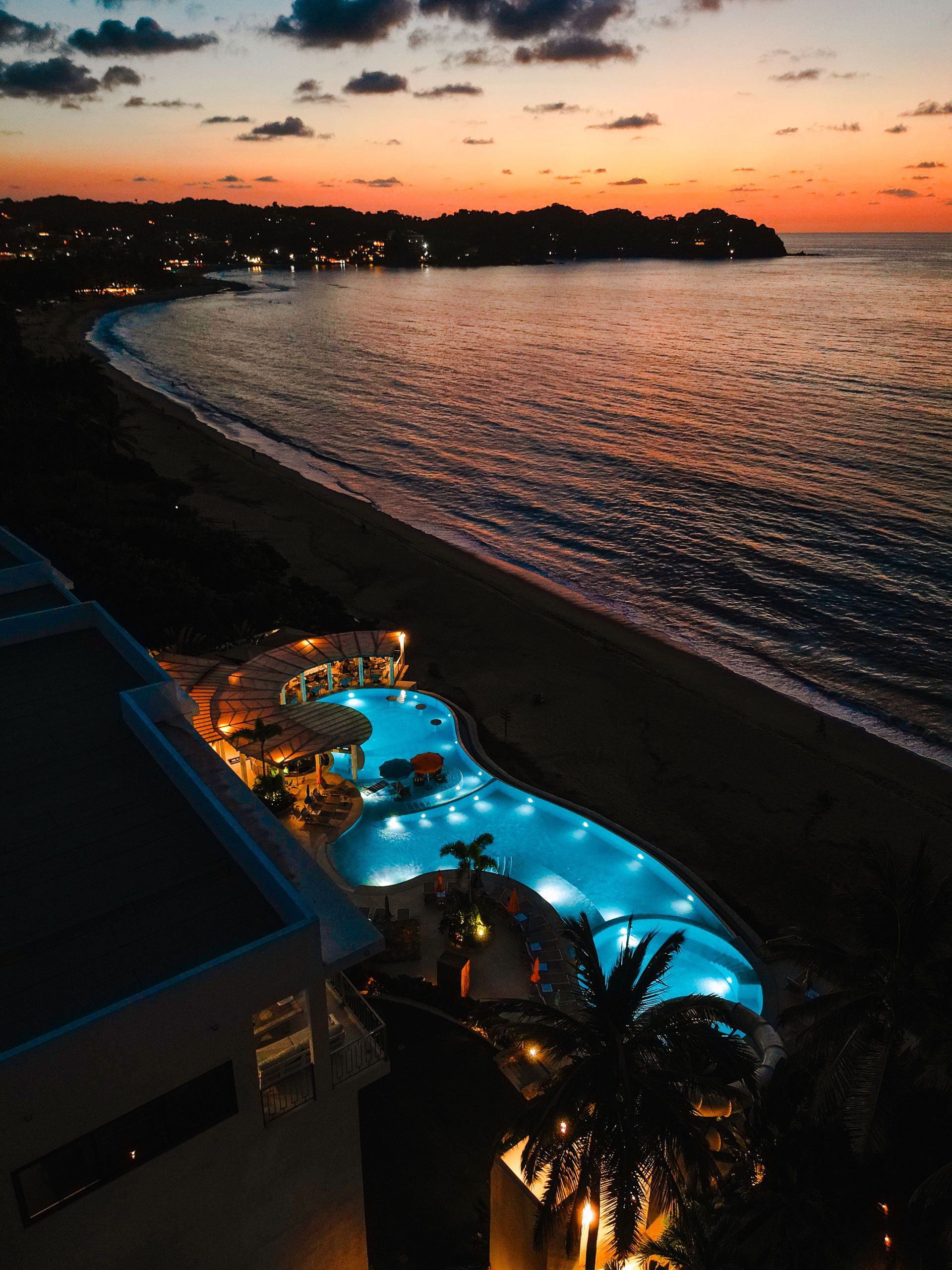 Sunset over a seaside resort with a glowing blue pool beside the shoreline