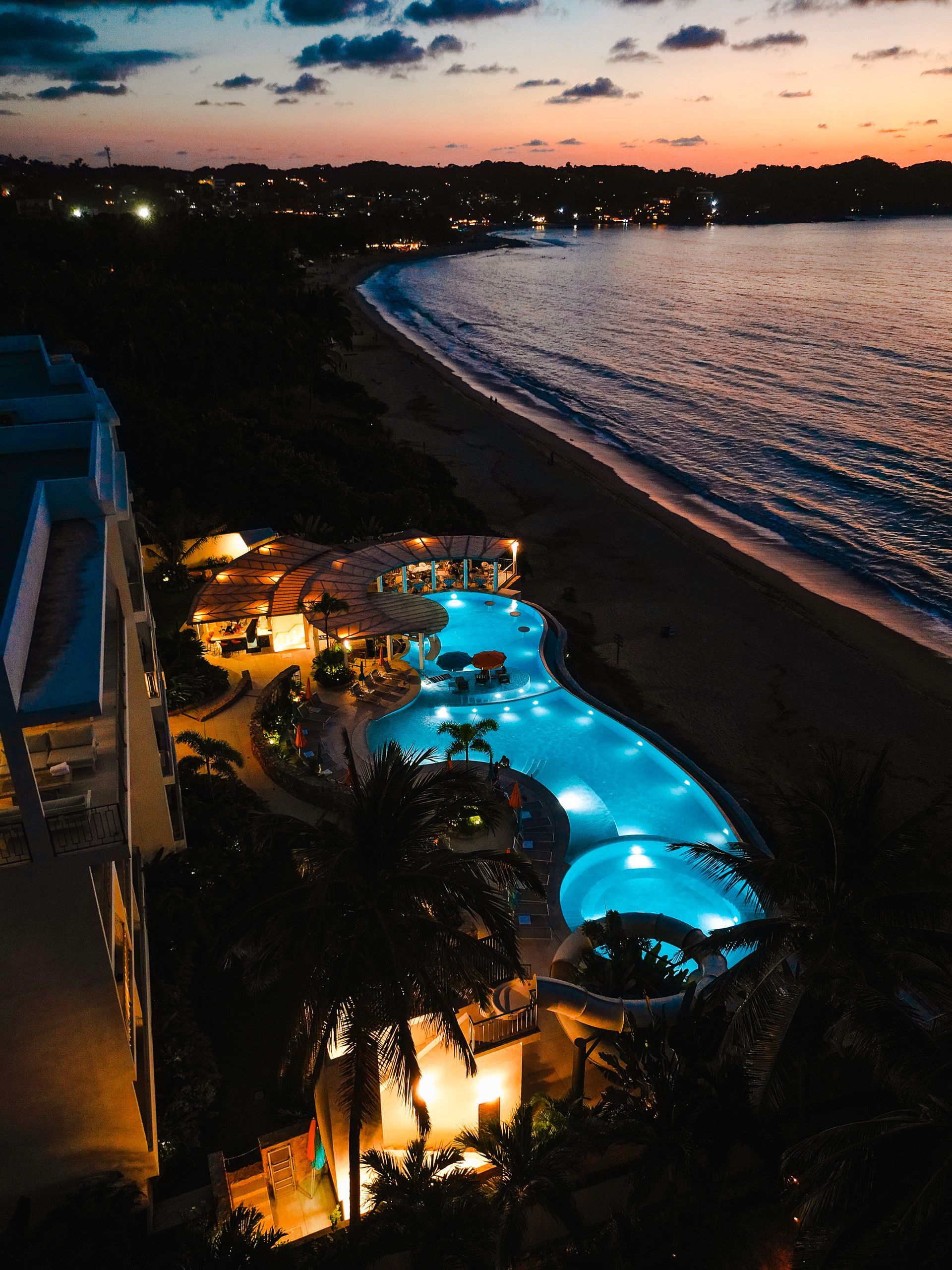 Beachfront resort pool glowing blue at dusk beside a dark shoreline and calm sea