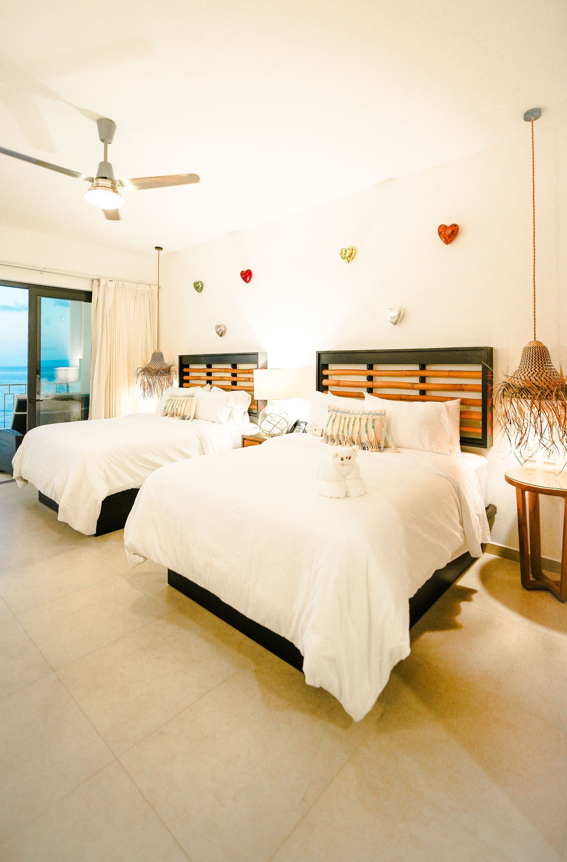 Bright bedroom with two white beds, wicker lamps, heart wall decor, and a ceiling fan