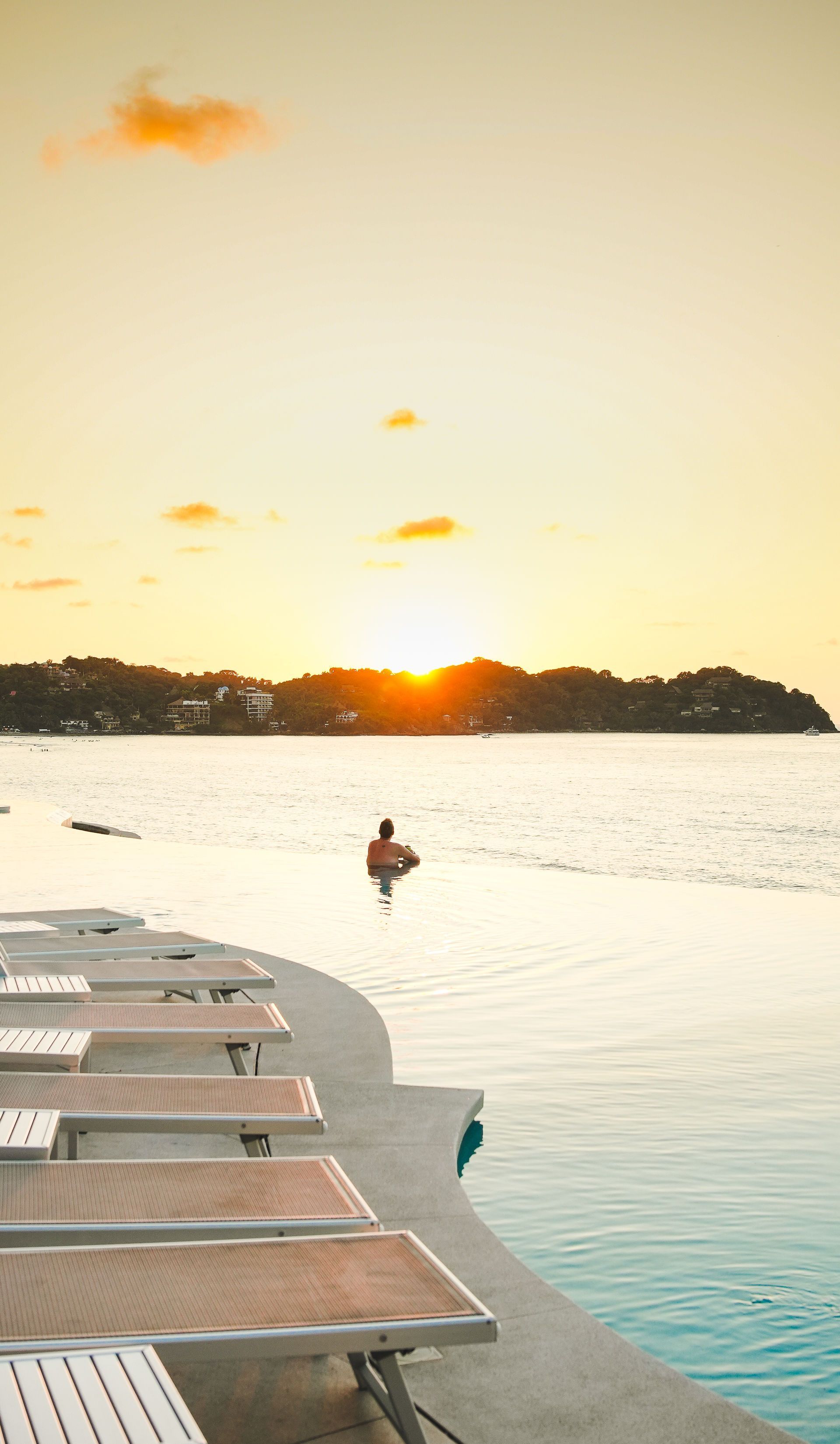 Sunset over a calm pool with lounge chairs on a deck and one swimmer near the horizon