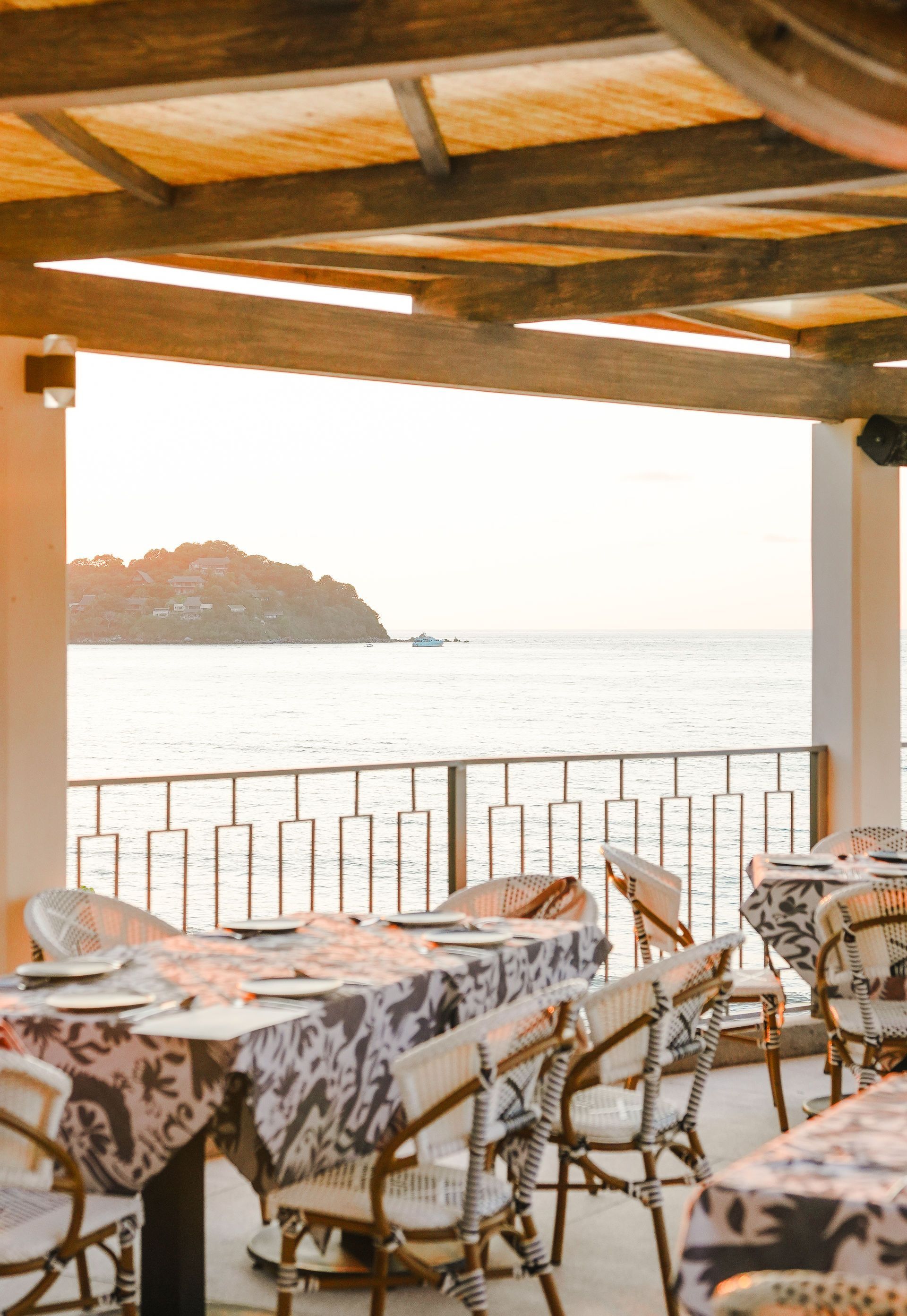 Seaside terrace with tables and chairs under a wooden roof, overlooking calm water at sunset