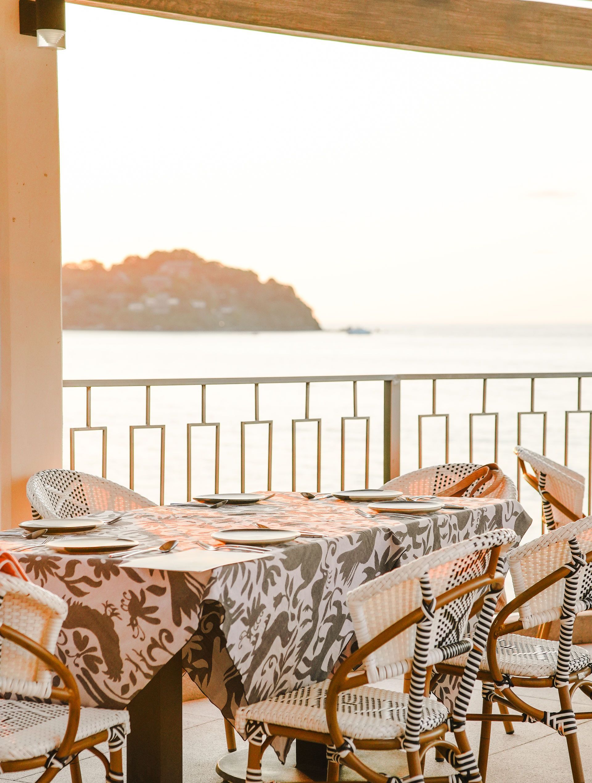 Sunlit seaside patio with wicker chairs and a patterned tablecloth overlooking calm water and a distant island