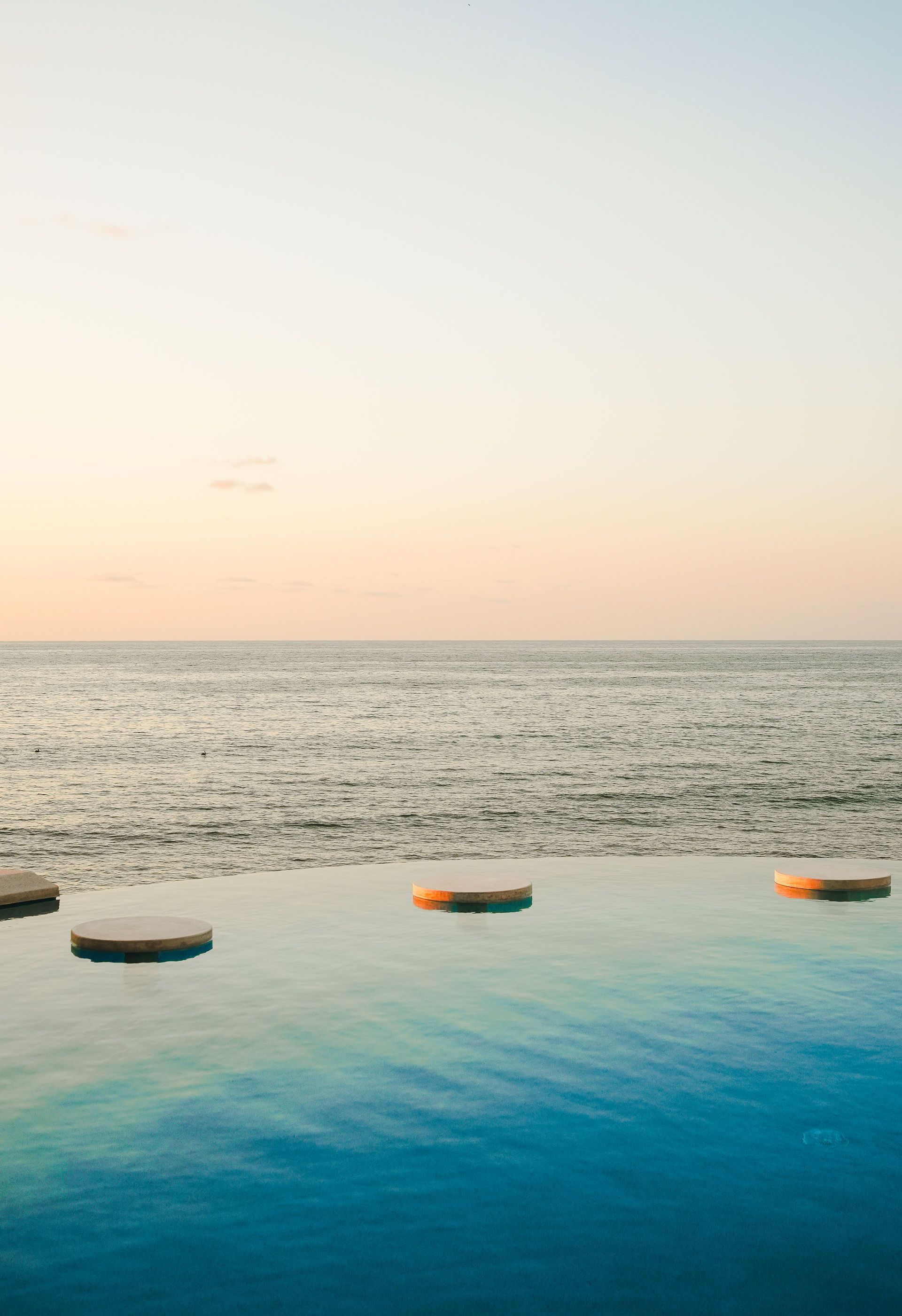Infinity pool overlooking a calm ocean at sunset with floating round loungers