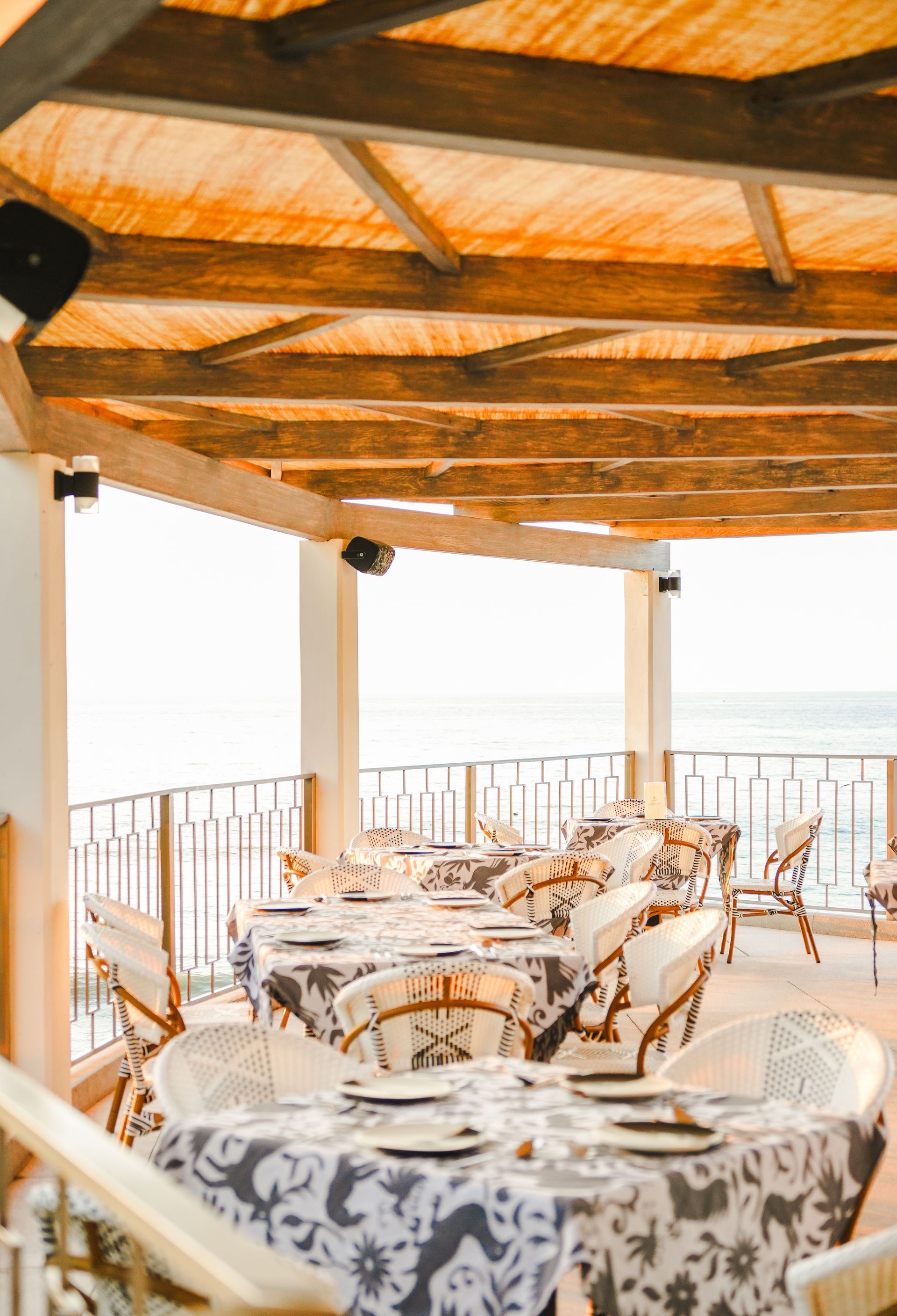 Sunlit seaside café terrace with patterned tables, wicker chairs, and wooden beams overlooking the water