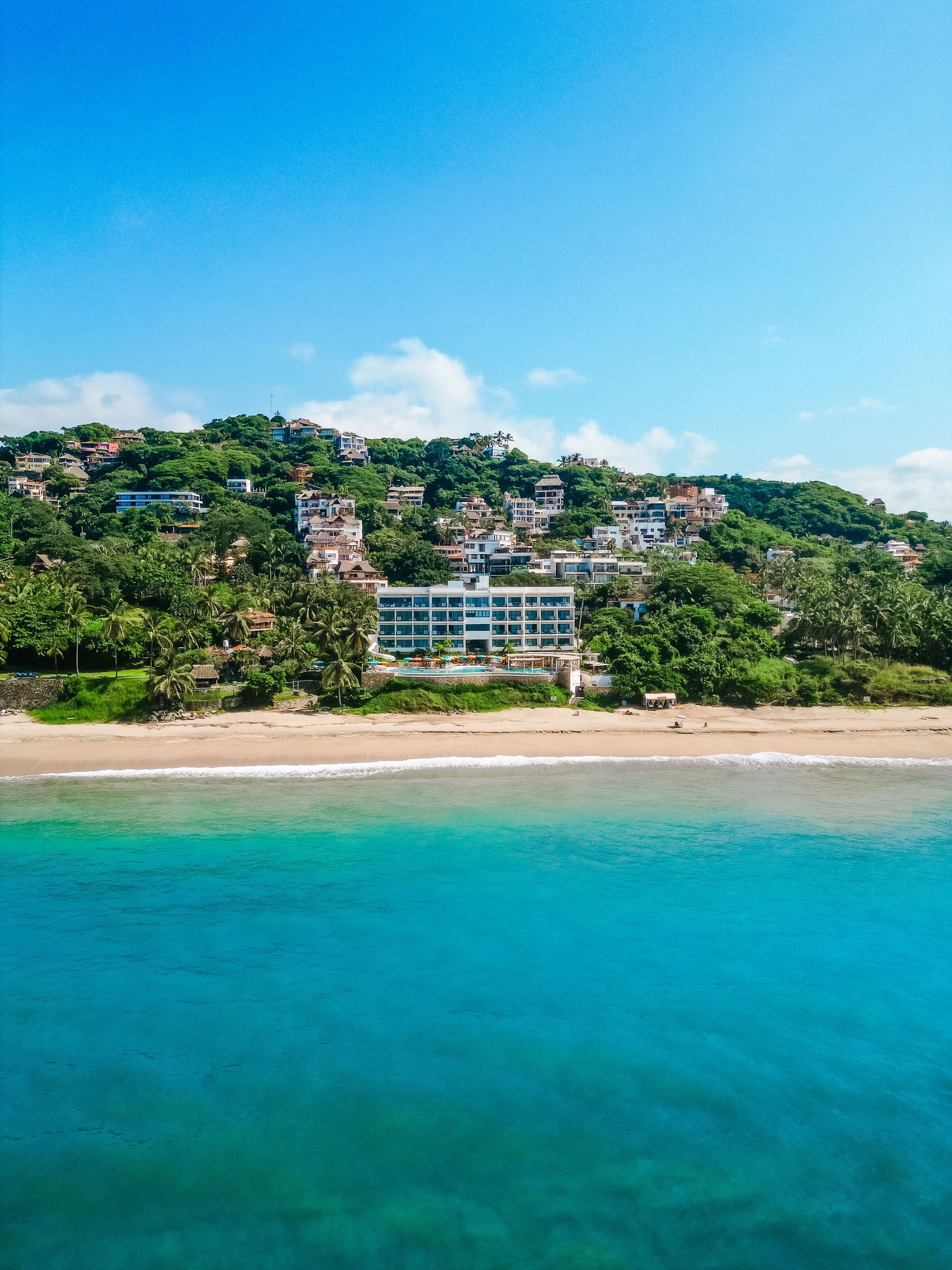 Tropical beachfront resort with turquoise water, sandy shore, and green hillside homes under a blue sky