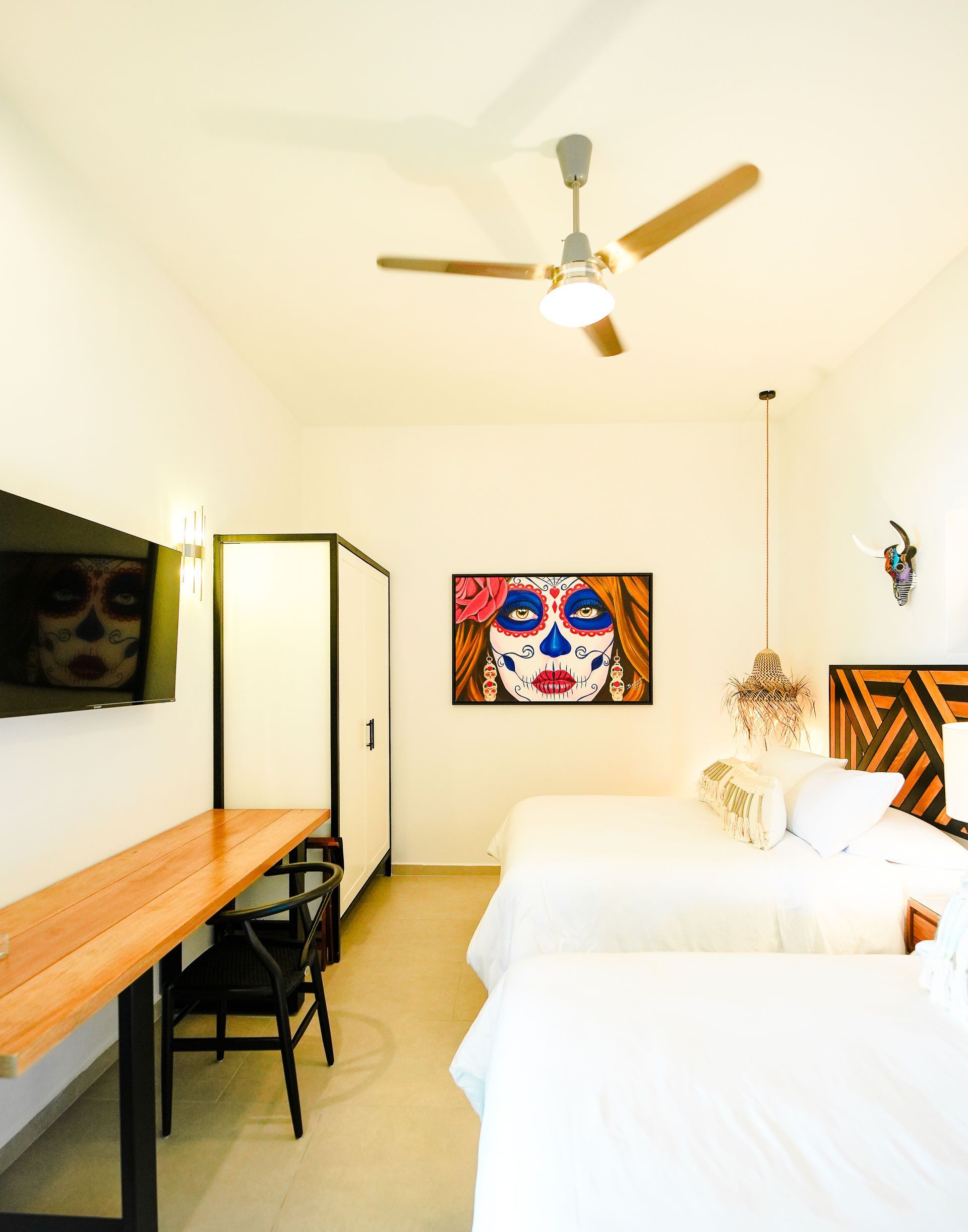 Bright bedroom with two white beds, wooden desk, wall art, and ceiling fan.