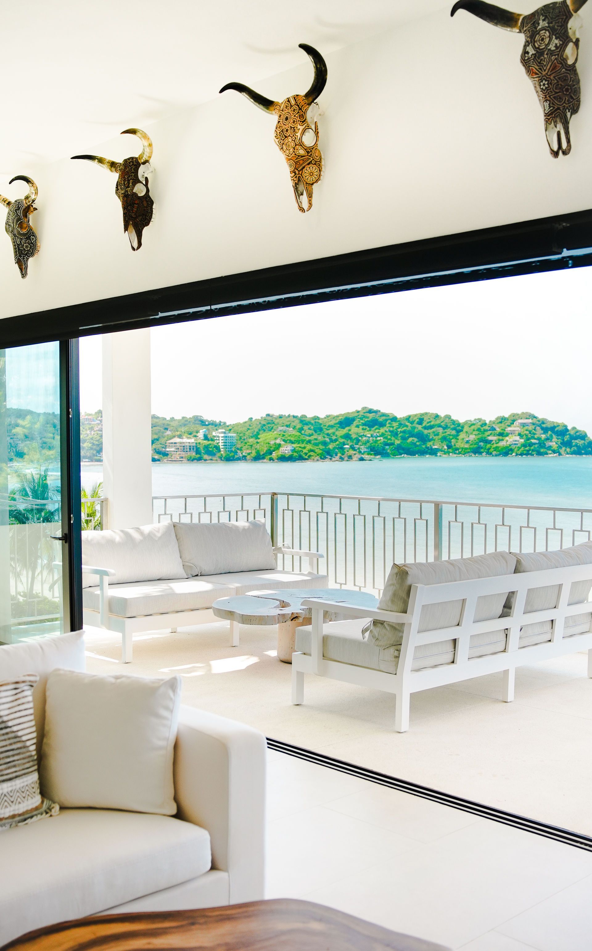 Bright waterfront patio with white lounge furniture, glass railing, and mounted antelope heads indoors.