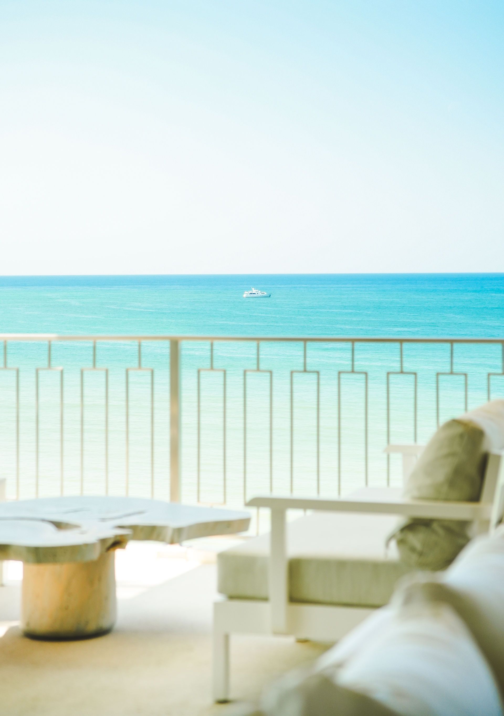 Bright seaside balcony with white chairs and table overlooking turquoise ocean and a distant boat