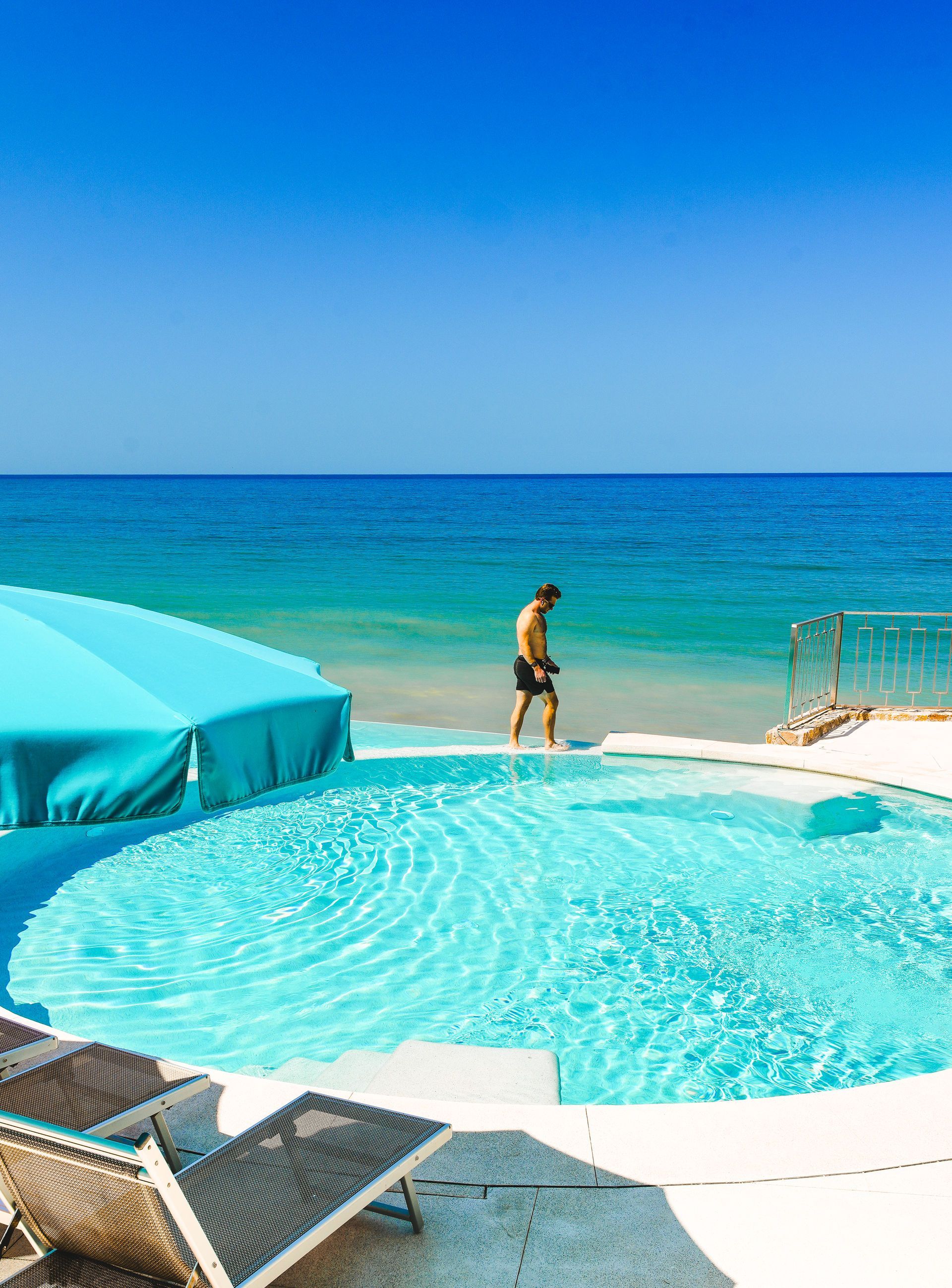 Oceanfront pool with turquoise water, lounge chairs, and a person walking by the beach