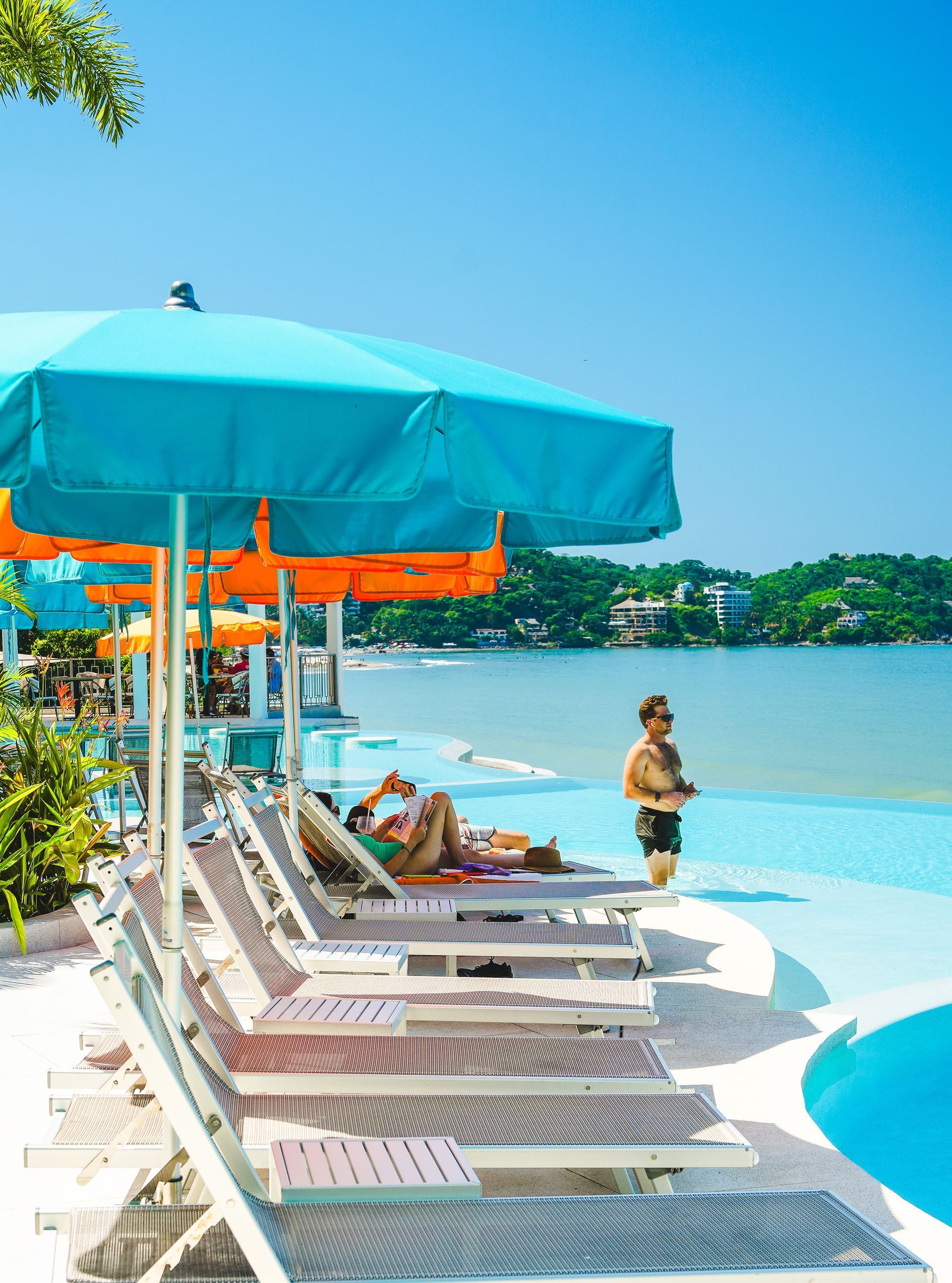 Turquoise umbrellas and lounge chairs beside a tropical beach pool with one person standing near the water