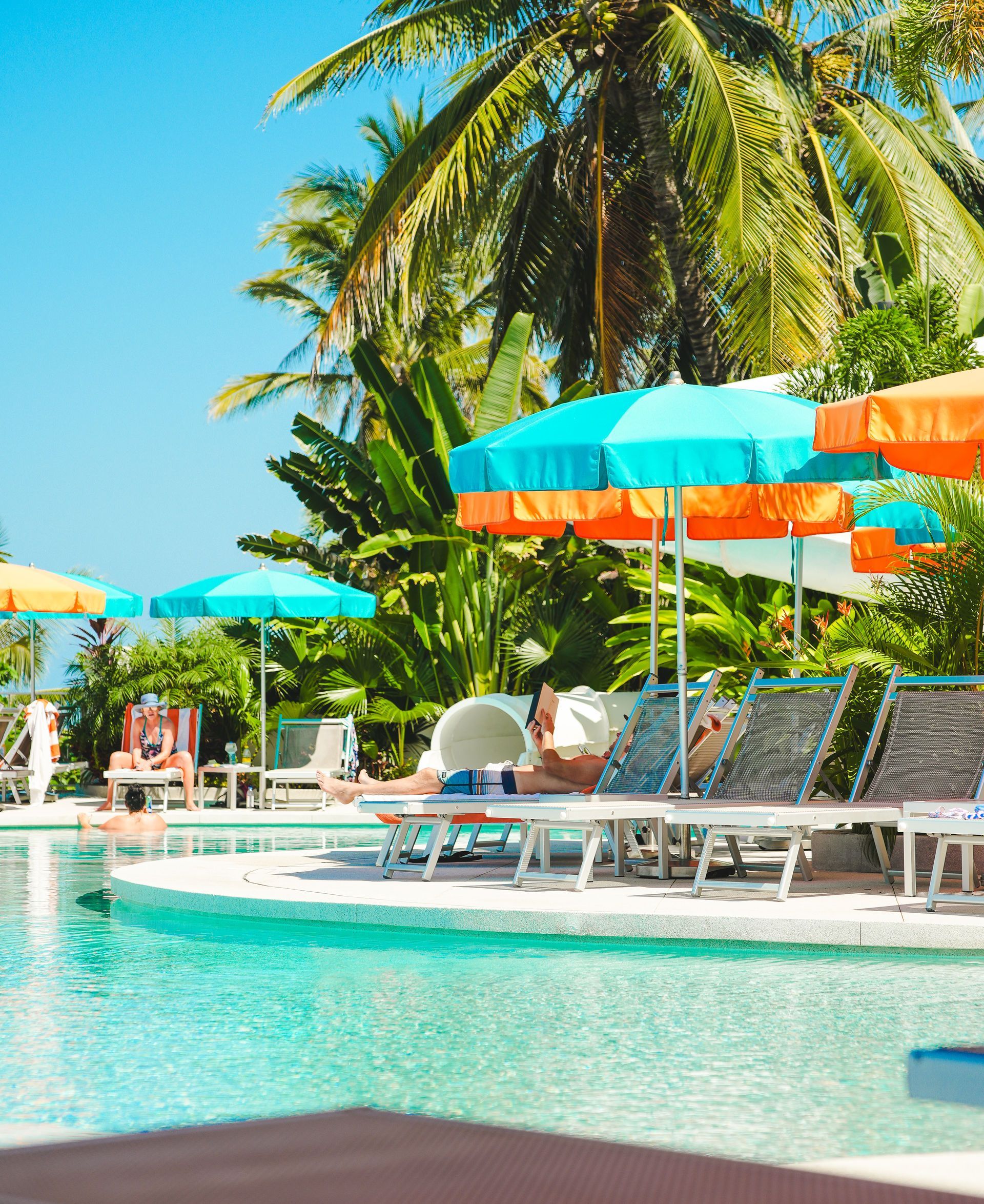 Tropical resort pool with white lounge chairs, colorful umbrellas, and palm trees under a bright blue sky