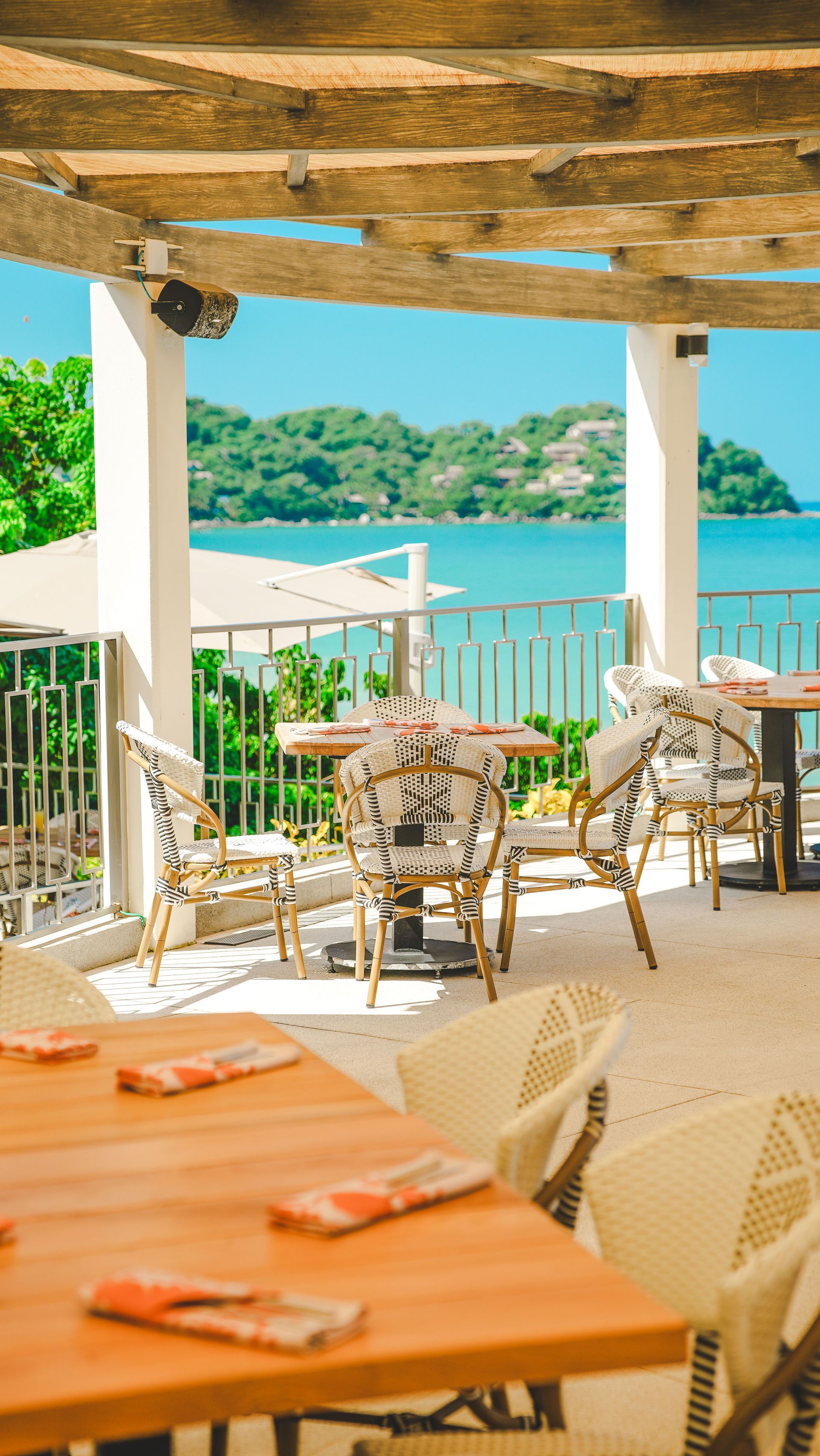 Open-air seaside café with wicker chairs, wooden tables, and turquoise water beyond a railing