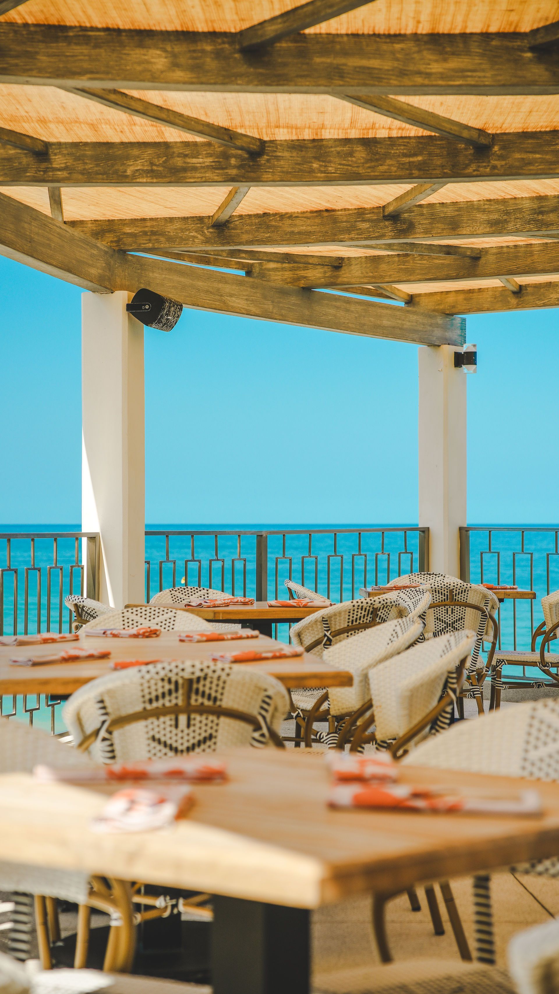 Shaded seaside café with wicker chairs, tables, and blue ocean view beyond the railing