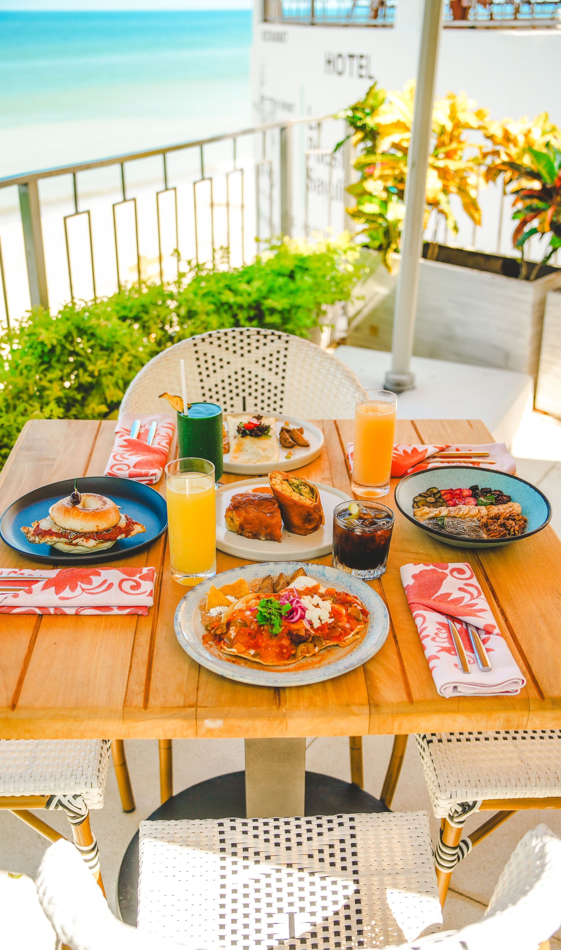 Sunlit outdoor table set with colorful breakfast plates, drinks, and flowers by the sea
