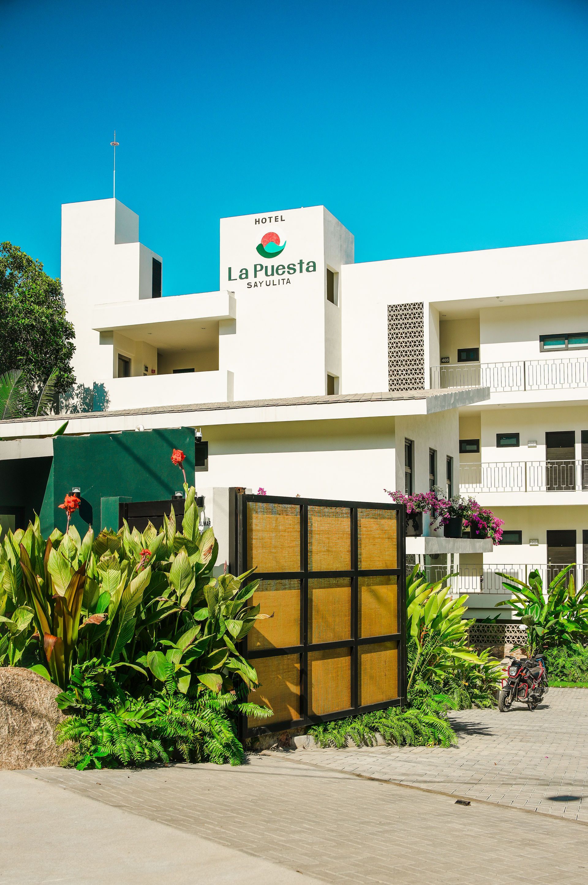Modern white building with turquoise sign and lush green plants under a bright blue sky