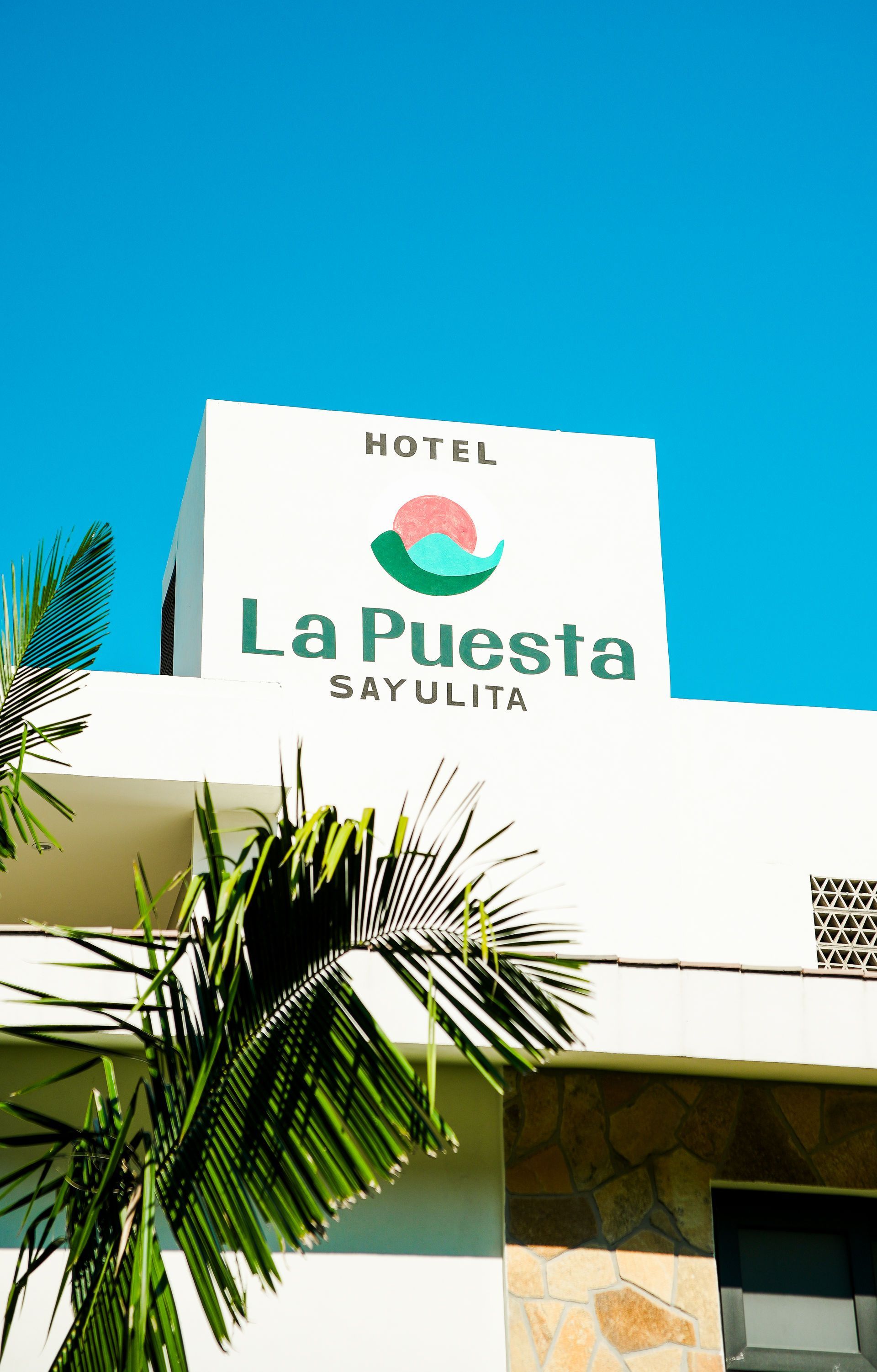 Hotel La Puesta sign on a white building against a bright blue sky, with palm leaves in front