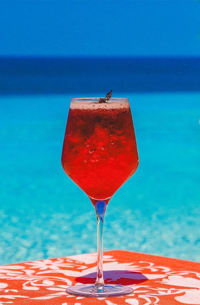 Red cocktail in a stemmed glass on an orange table by a turquoise pool and blue sea.