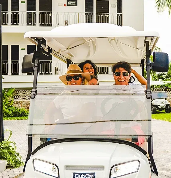 Three people smiling in a white golf cart outdoors, wearing sunglasses and a straw hat.