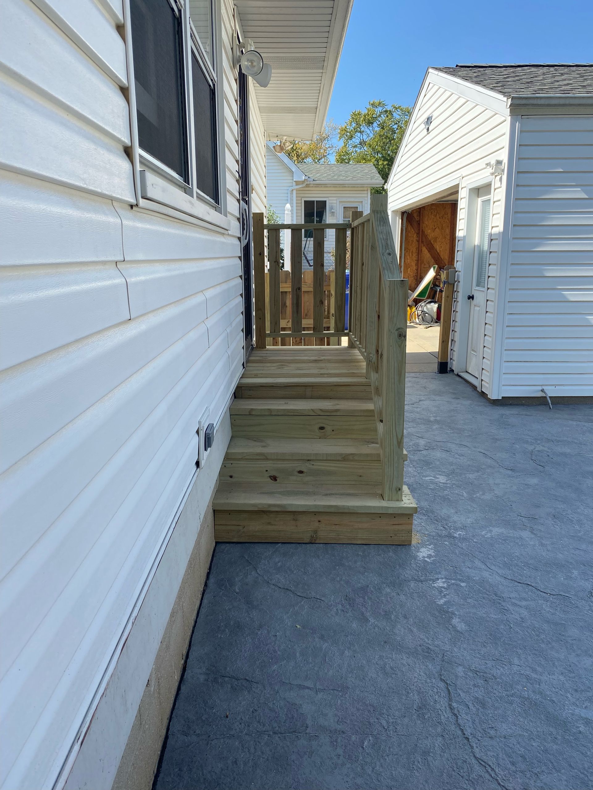 Wooden porch steps beside a white-sided house, leading to a small landing and gate near a detached shed.