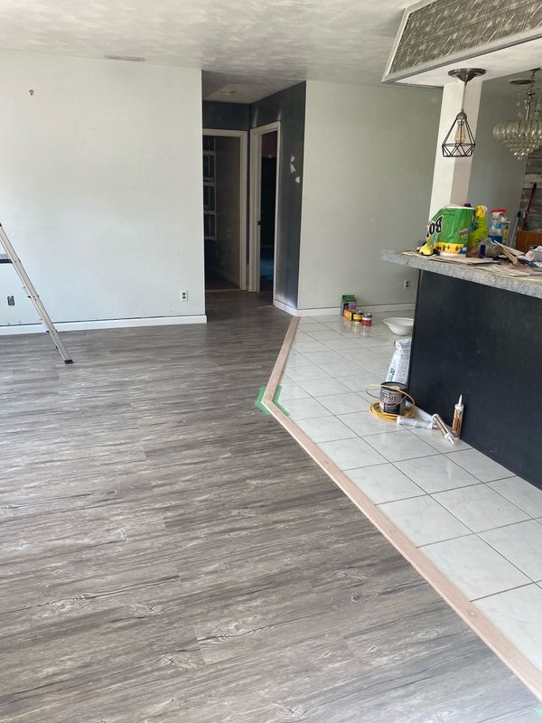 Empty modern living room with gray floors, white walls, and a dark kitchen counter on the right.