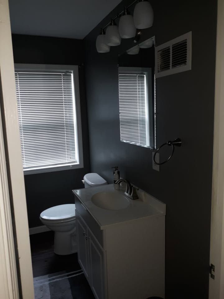 Small dark bathroom with white vanity, toilet, sink, mirror, windows, and shower curtain rod
