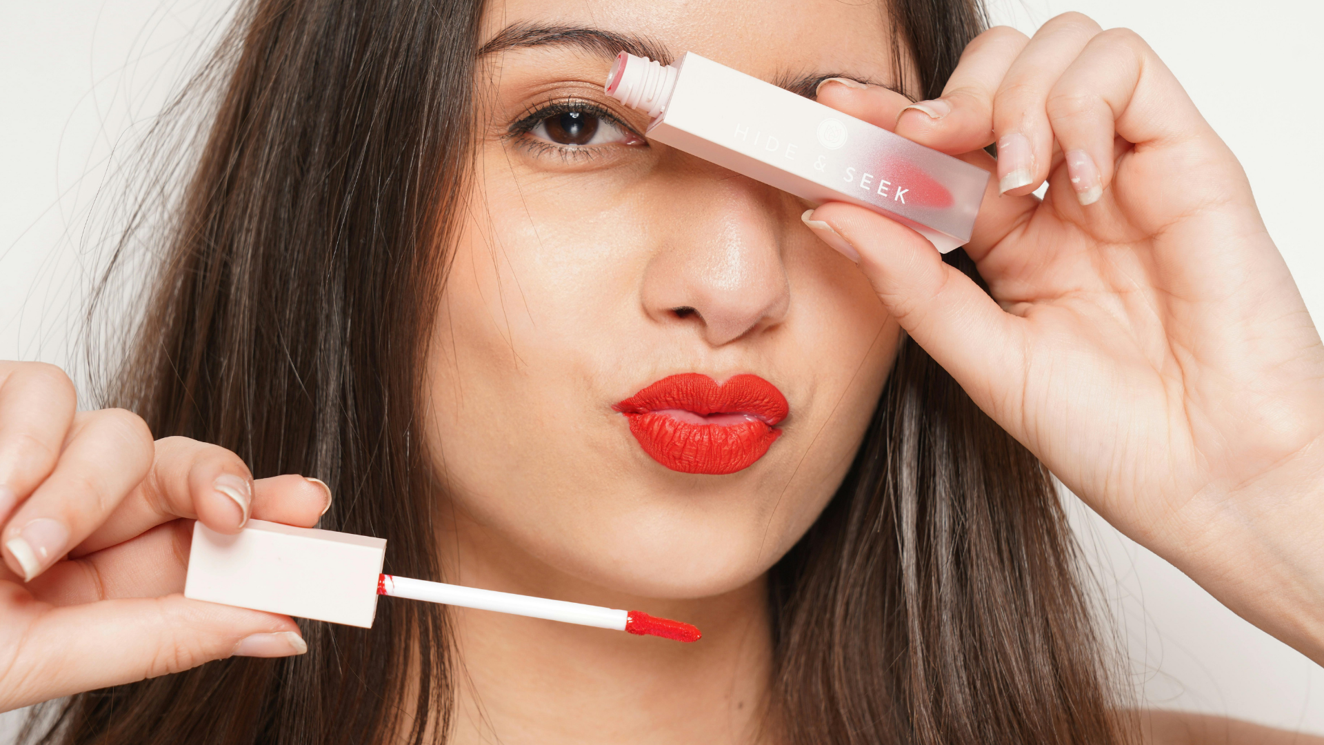 Woman holding red lipstick wand near her face, applying bright red lipstick against a white background