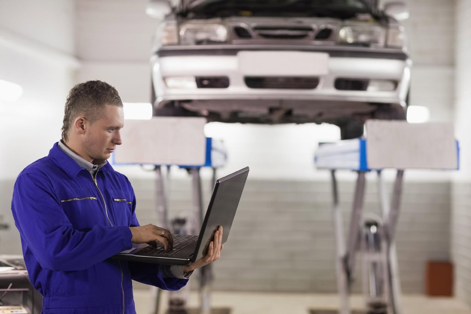 Mechanic in blue coveralls using a laptop beside a car on a lift in an auto shop | Frontier Auto Center