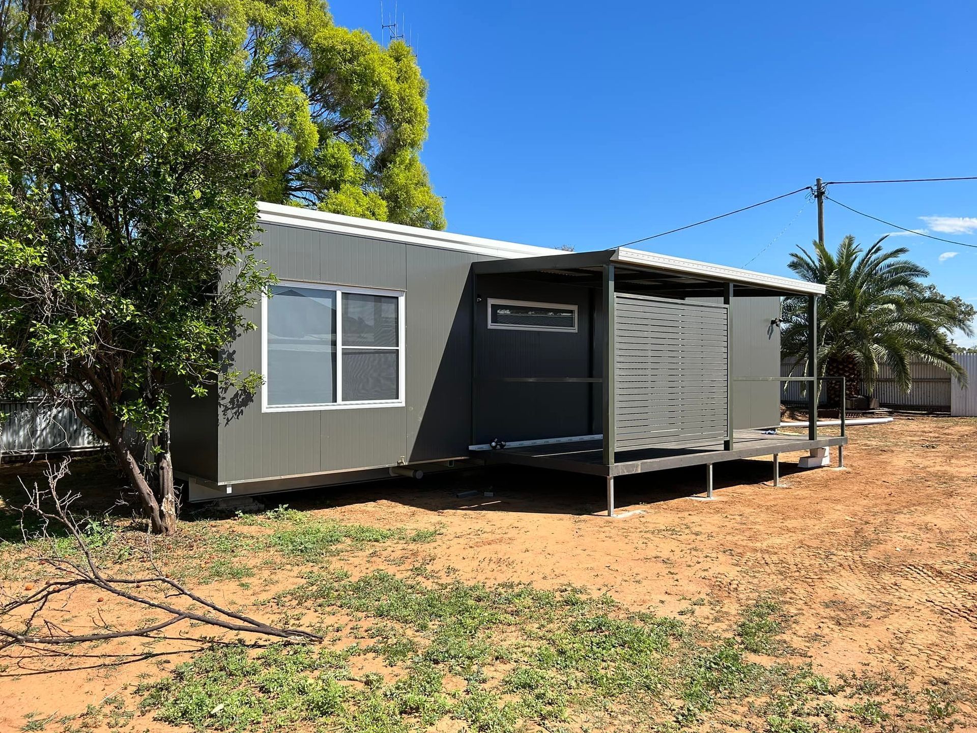 A Modern, Dark-gray Rectangular Cabin With a Covered Wooden Deck Stands in a Dry — OSM Transportables In Dubbo, NSW